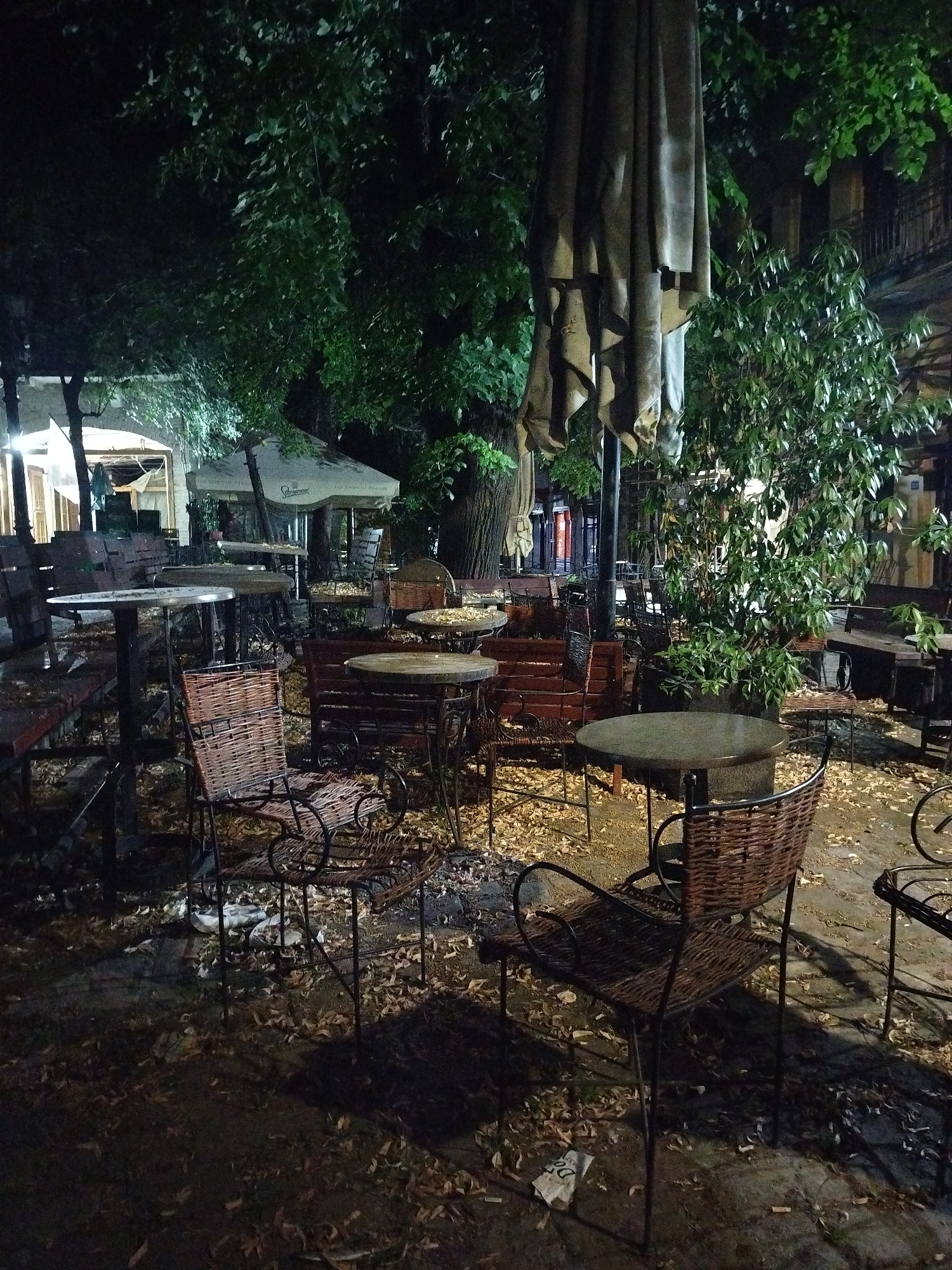 The image depicts an outdoor seating area of a cafe or restaurant at night. The scene is dimly lit, with artificial lights illuminating some of the tables and chairs. There are numerous tables and chairs scattered throughout the area, many of which appear to be made of wood and wicker. Some tables are round with metal bases, while others are rectangular. Large umbrellas, likely for shade during the day, are folded and secured, one of which is very prominent in the center of the image. Green foliage from trees overhangs the seating area, and fallen leaves are visible on the ground, suggesting it may be autumn. In the background, there are hints of buildings, possibly part of the establishment or adjacent structures. The atmosphere is quiet and deserted, with no people present. The general appearance suggests an establishment in a part of Novi Sad, Serbia, that is likely a popular outdoor gathering spot.