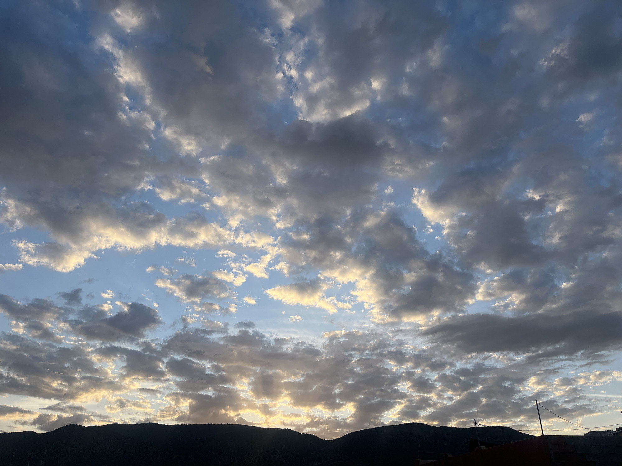 The image depicts a cloudy sky over Ouaouizert, Morocco. The sky is filled with altocumulus clouds, some of which are catching the light of the setting sun. Below the clouds, in the foreground, is the silhouette of a mountain range. A few buildings are visible at the base of the mountains. The overall light suggests it is either sunrise or sunset. There is no evidence of people or interaction.