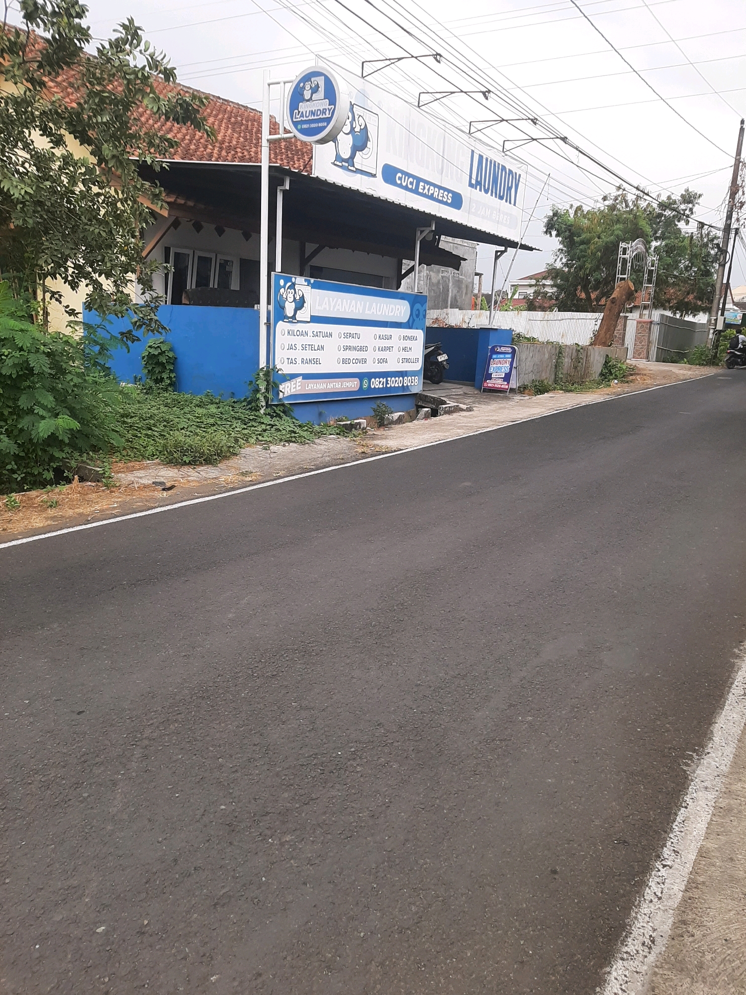 In Sumampir, Indonesia, a laundry service business is situated in a low-rise building with a blue facade. The establishment features prominent signage, including a circular logo with a cartoon gorilla and the text "KIRIM KONG LAUNDRY" and "CUCI EXPRESS," along with smaller text indicating "2 JAM BERES." Below this, a blue sign details services such as "KILOAN SATUAN," "JAS, SETELAN," "TAS, RANGSEL," "KASUR," "BONEKA," "SPRINGBED," "BED COVER," "SOFA," "HELM," and "STROLLER." The sign also advertises "LAYANAN ANTAR JEMPUT" (pick-up and delivery service) with the phone number 0821 3020 8038. The street in front of the laundry is asphalted, with a white line marking the edge of the road. Dense green foliage grows along the side of the road, partially obscuring a concrete drainage channel. The sky is overcast.