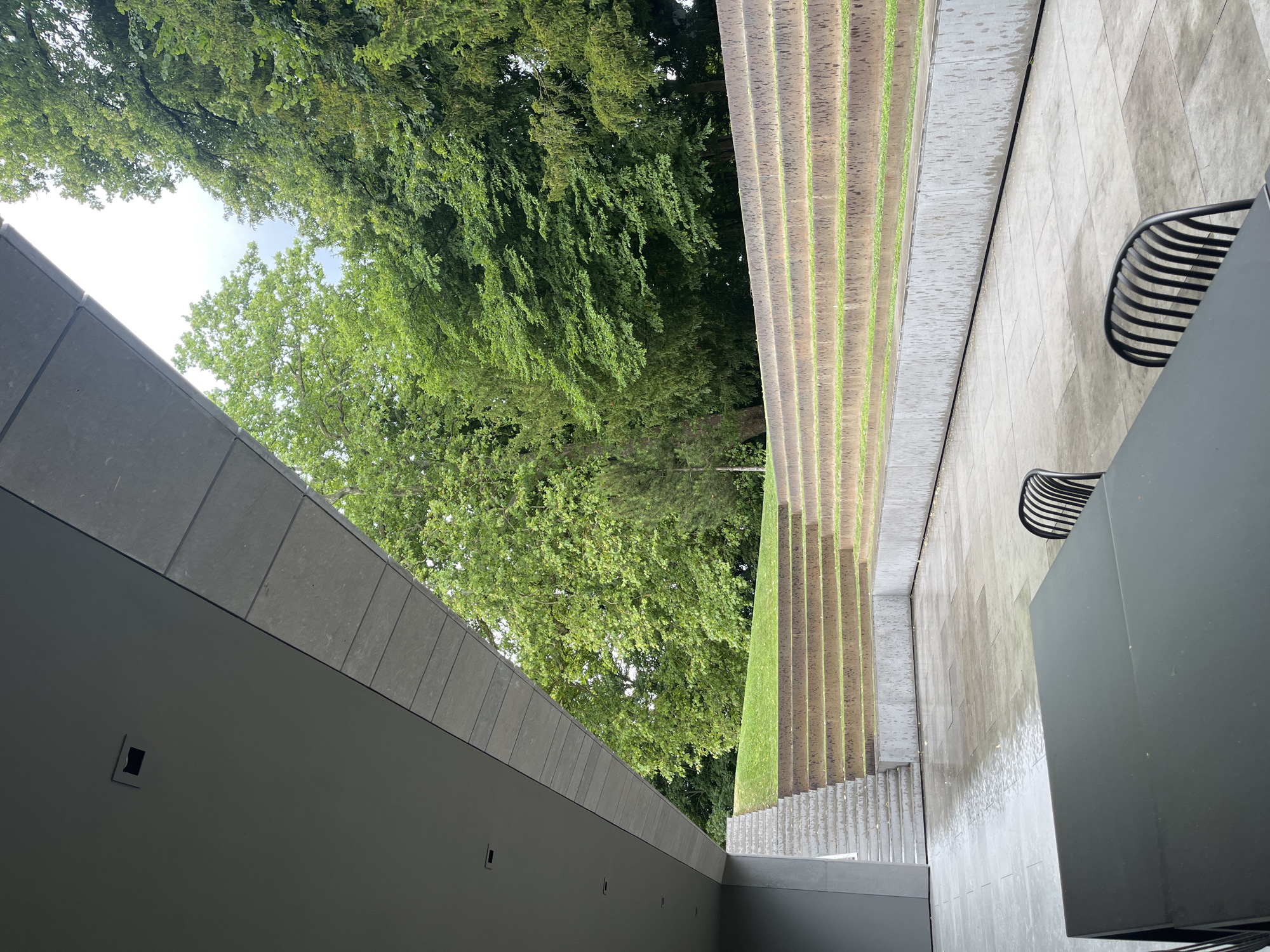 The image depicts an outdoor area with modern architecture and natural elements. In the foreground, a black rectangular table with two black chairs are visible on a wet, grey tiled patio. To the left, a grey, angular overhang of a building extends into the frame. Beyond the patio, a tiered seating area made of stone with grass growing in the crevices ascends towards a grassy slope. This seating area is a prominent feature of the landscape. In the background, lush green trees form a dense canopy against a cloudy sky. The scene appears to be a quiet outdoor space, possibly a public area or a private residence, located in Cologny, Switzerland, suggested by the surrounding greenery and the architectural style often found in the region.