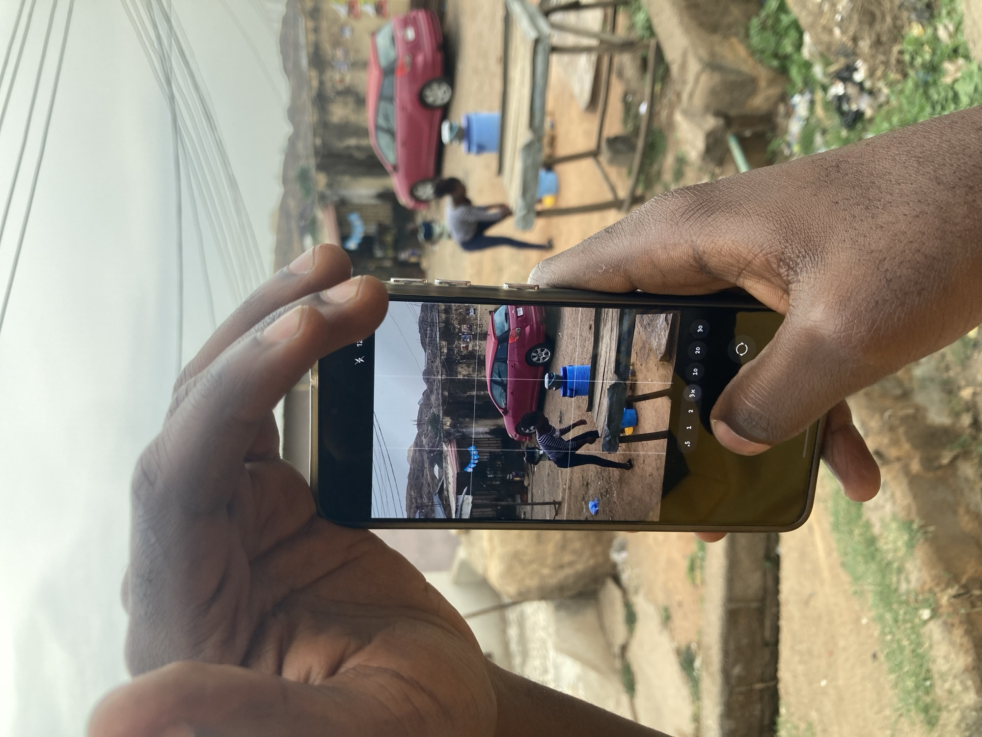 A person is holding a smartphone to take a photograph of a scene in Jos, Nigeria. The phone's screen shows a red car, a person sitting near a blue water container, and some buildings, possibly houses or shops. The foreground includes the person's hands holding the phone, and the background shows a slightly blurred view of the same scene. The environment appears to be a populated area with buildings and structures.