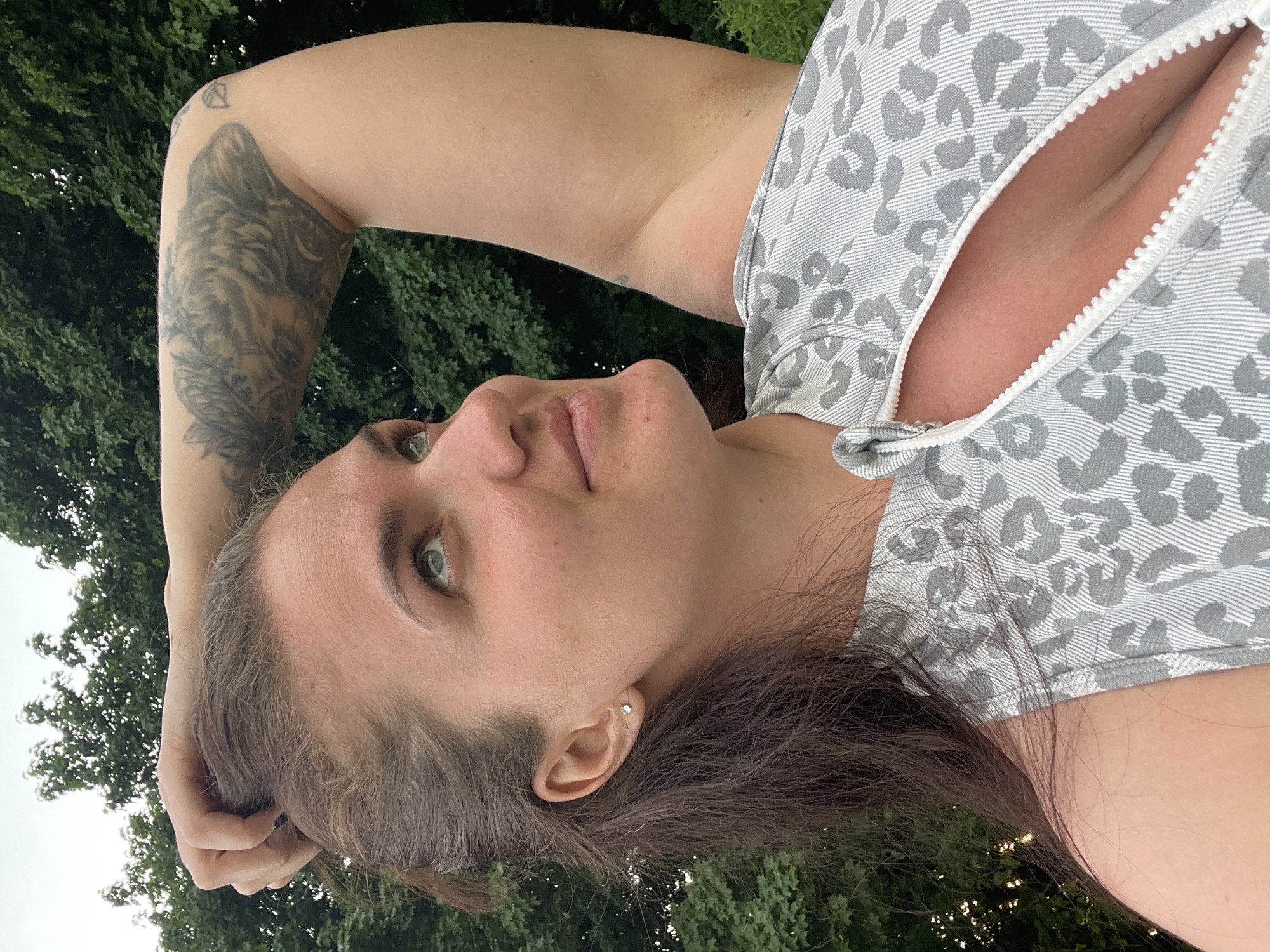 A woman lies on the grass. The woman has light skin, dark hair, and green eyes. She wears a light gray and white patterned top with a zipper. Her right arm is raised, with a large tattoo visible on her upper arm. The background shows green trees. The location in the city of Ostrov, Russia is not discernible.