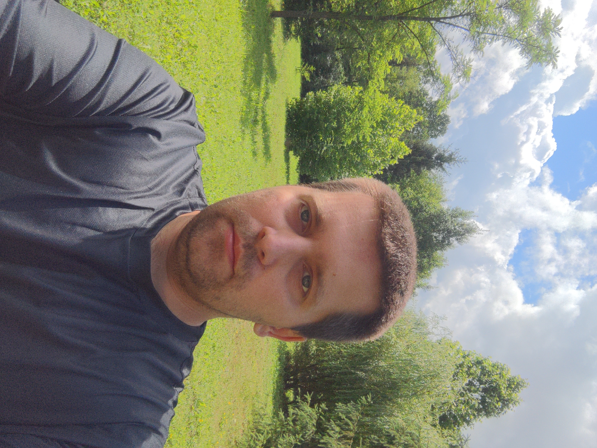 A man is taking a selfie outdoors in a park-like setting. He is positioned in the foreground, facing the camera. He has short brown hair and is wearing a dark grey t-shirt. The background is filled with lush green trees, grass, and a bright blue sky with white clouds. The location appears to be a park or a large garden, with a well-maintained lawn and various types of trees, including some with weeping branches on the right. The overall impression is one of a sunny day in a natural environment. No specific landmarks or buildings are visible to pinpoint the exact location within Holubyne, Ukraine, beyond it being an outdoor, green space.