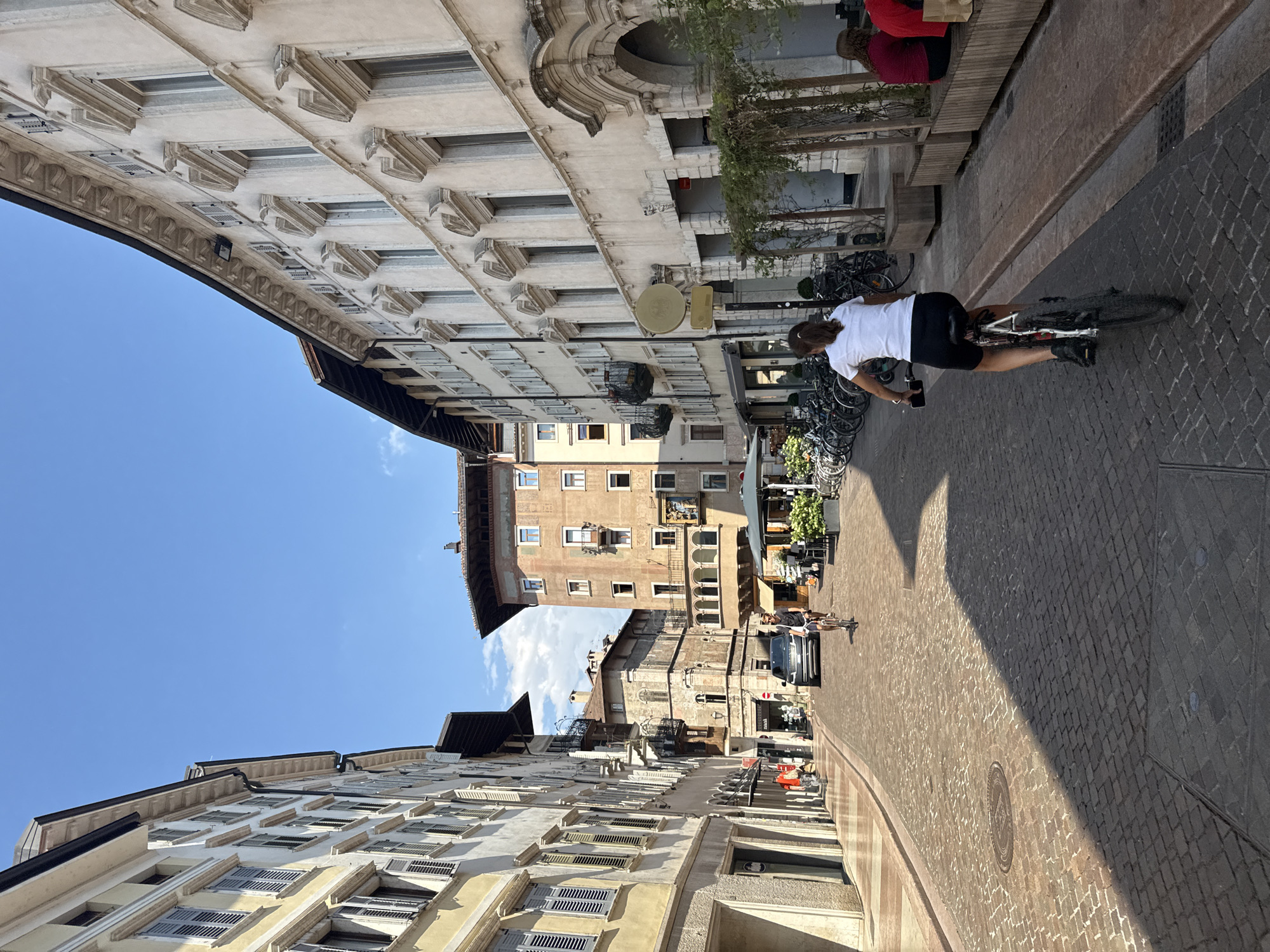 A street scene in Trento, Italy, captures a woman cycling towards the viewer down a cobblestone street lined with ornate buildings. The woman, wearing a white t-shirt and black shorts, is riding a bicycle with a black frame and silver handlebars. She is positioned in the foreground, dominating the lower right quadrant of the image. Behind her, a man on a bicycle is also heading away from the viewer, and a dark-colored SUV is parked further down the street.
The architecture on both sides of the street features tall buildings with multiple windows, some adorned with shutters. The buildings on the left are a lighter yellow hue, while those on the right are a creamy white. The street is paved with cobblestones, and a clear shadow cuts diagonally across it, indicating a sunny day. Bicycles are parked in racks along the side of the street on the right, and outdoor seating areas for cafes are visible. The sky is a clear, bright blue with a few scattered clouds. The overall impression is of a vibrant, pedestrian-friendly urban environment in Italy.
