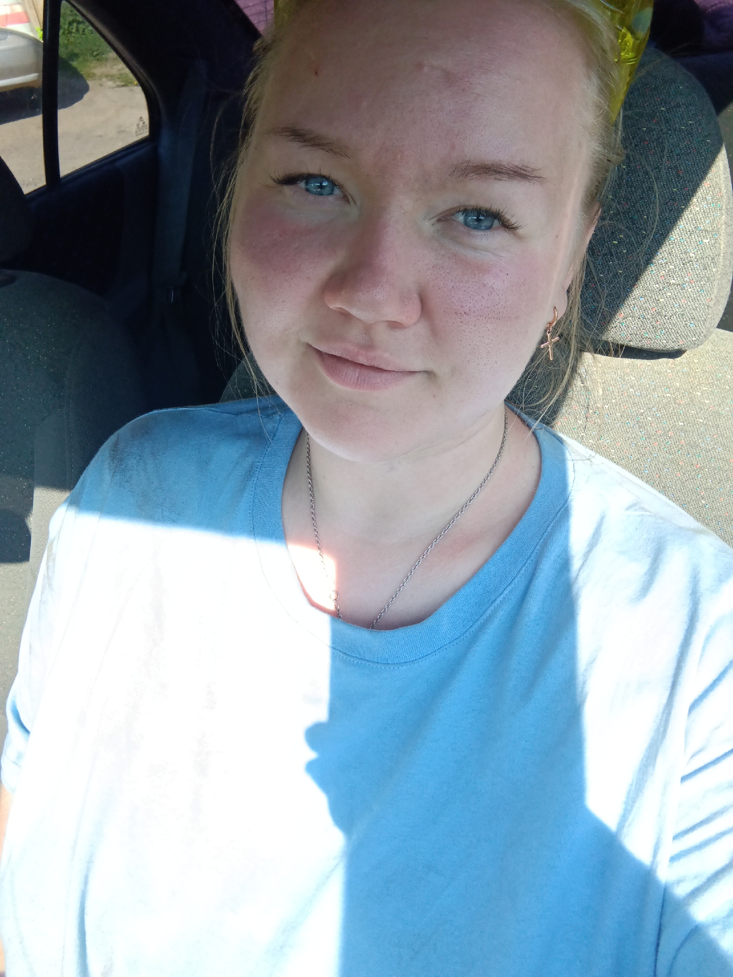 A Caucasian woman with fair skin, light blonde hair, and blue eyes sits inside a car. She is wearing a light blue t-shirt and a silver chain necklace. She has small gold cross earrings. The interior of the car includes a grey fabric seat and a dark window in the background. Light is shining on her shirt. The specific location of the city of Фоки, Russia, cannot be determined from the image.