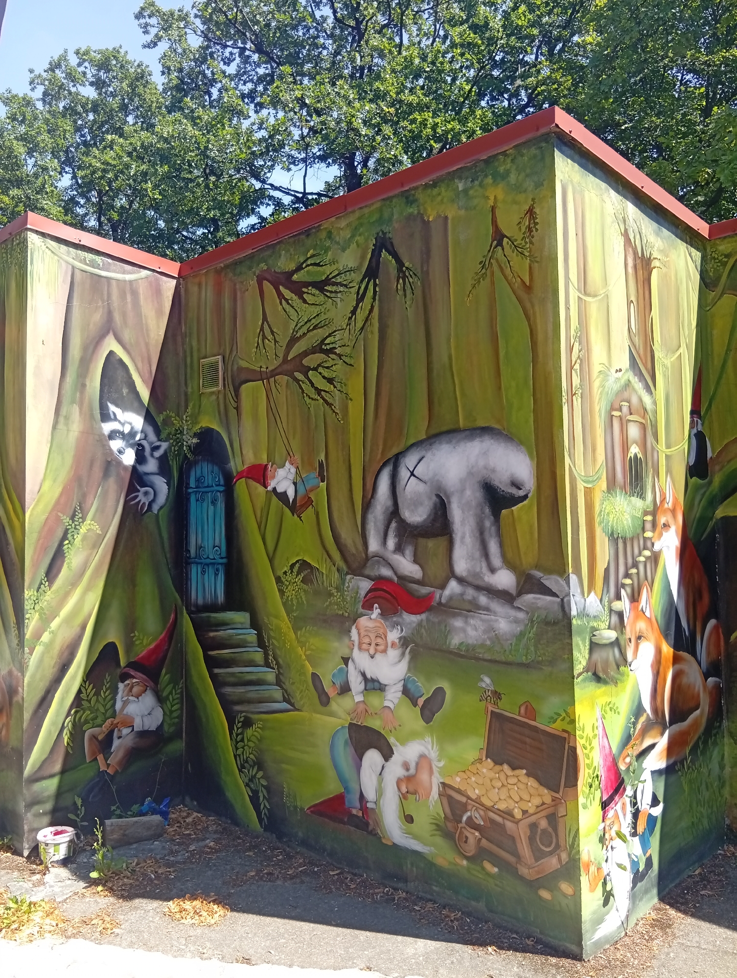 This image depicts a vibrant mural painted on the exterior walls of a building in the city of Sobótka, Poland. The mural portrays a whimsical forest scene filled with fantasy creatures. On the left side, a raccoon peers out from a hollow tree trunk. A blue door with a step leads into the tree. A gnome with a red hat is seated and playing a flute next to the steps. Another gnome is depicted swinging on a rope swing from a tree branch. In the center, a large, gray, bear-like figure with a cross marked on its back stands on rocks. To the right, two foxes are positioned near a stone structure resembling a small castle or tower. Several other gnomes are scattered throughout the scene, including one sitting on the ground, another lying down near a treasure chest filled with gold coins, and a final one peeking from behind the corner of the building. The overall environment is lush and green, suggesting a forest setting, with trees and foliage dominating the background. The building itself appears to be a small structure, possibly a public facility, with a red roofline visible at the top. The ground in front of the mural is paved and covered with fallen leaves and some scattered debris.