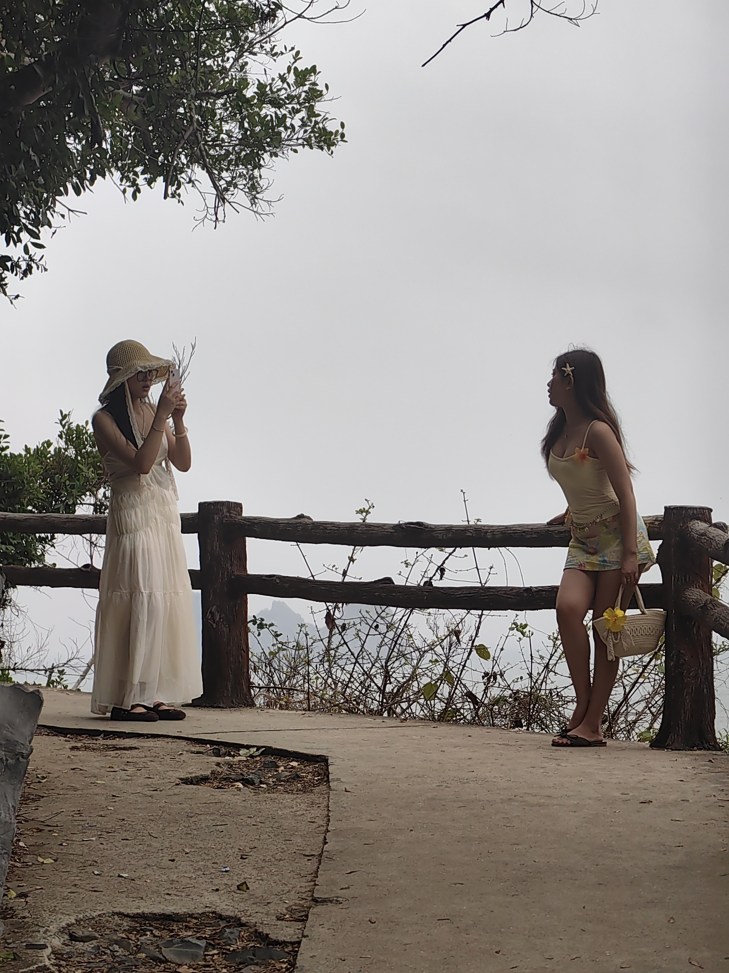 Two young women are on an elevated concrete pathway overlooking a landscape obscured by mist or clouds. A wooden railing surrounds the pathway. One woman, wearing a long white dress and a straw hat, is taking a photograph with her phone. The second woman, wearing a yellow top and patterned shorts, leans against the railing, gazing into the distance. The location is in the city of Hải Phòng, Vietnam. The pathway shows signs of wear and debris. Foliage and branches are visible on either side of the pathway.