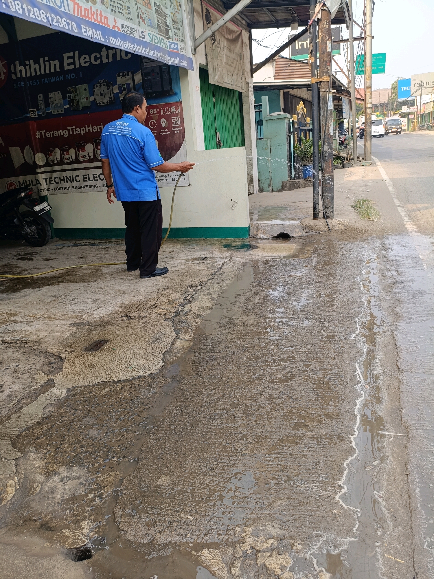 In Tangerang, Indonesia, a man wearing a blue shirt and black pants stands on a concrete surface, holding a hose that is spraying water. The water is creating puddles and running down the street. In the background, there is a building with a sign that reads "Shilin Electric". There is also a motorcycle parked to the left of the man. The scene appears to be a street in a commercial area.