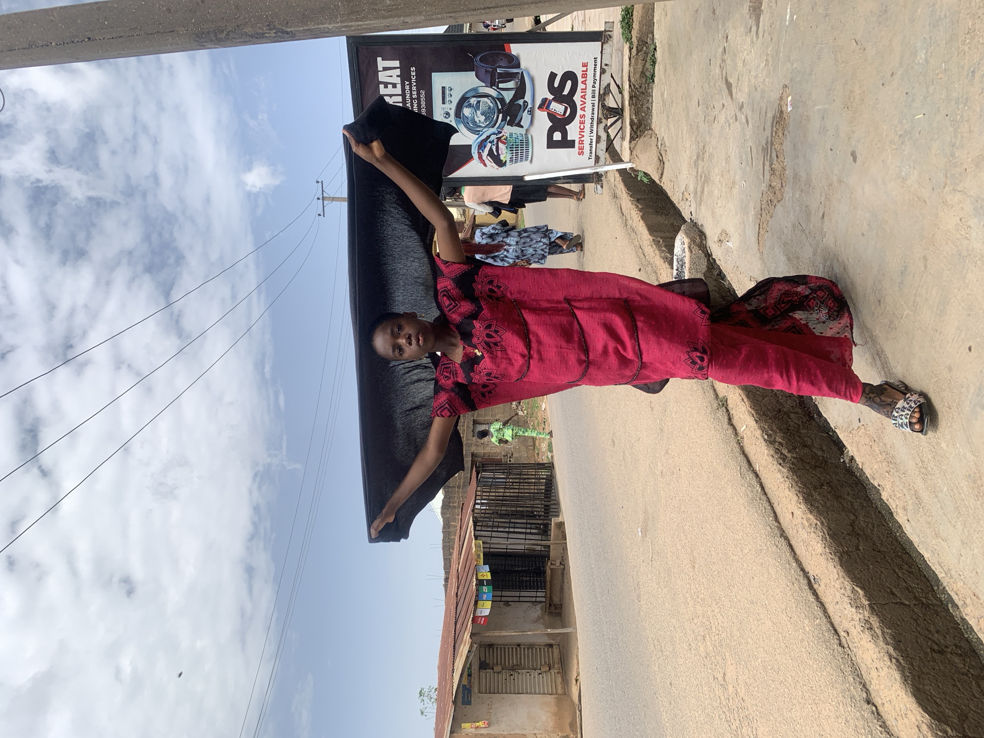A woman stands on a street in what is likely a residential or commercial area of Jos, Nigeria, holding a large, dark-colored piece of fabric outstretched in her hands. She is wearing a red, patterned outfit. Behind her, there's a building with a corrugated metal roof and a black iron gate. A sign is visible, advertising laundry and financial services (POS). Other people can be seen walking in the background. The sky is cloudy.