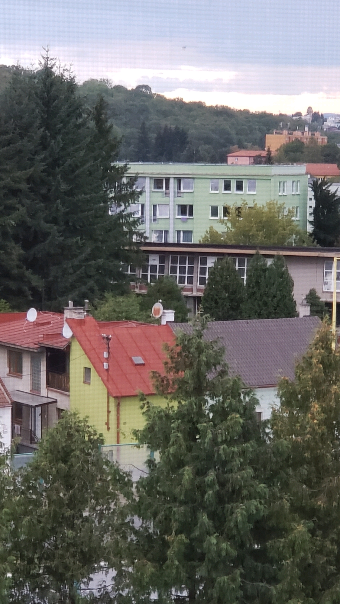 This is a daytime, eye-level, outdoor shot of a residential area, possibly an apartment complex and some houses. The scene is dominated by trees and buildings. In the foreground, several houses with red and green roofs are visible, interspersed with dense green foliage. One house has a bright yellow-green facade and a red roof. The middle ground features a large, light green apartment building with numerous windows. Behind this, the landscape rises to a wooded hill. The sky is overcast with hints of pink and orange, suggesting either dawn or dusk. A small, dark object, possibly a drone or a bird, is visible in the upper sky. The area appears to be residential, with satellite dishes on the roofs indicating modern amenities. There are no people visible in the image. The overall impression is of a suburban or semi-urban setting. Based on the architecture and the presence of the apartment complex, this location could be part of the Sekčov housing estate in Prešov, Slovakia, which is known for its numerous apartment buildings and surrounding green areas.