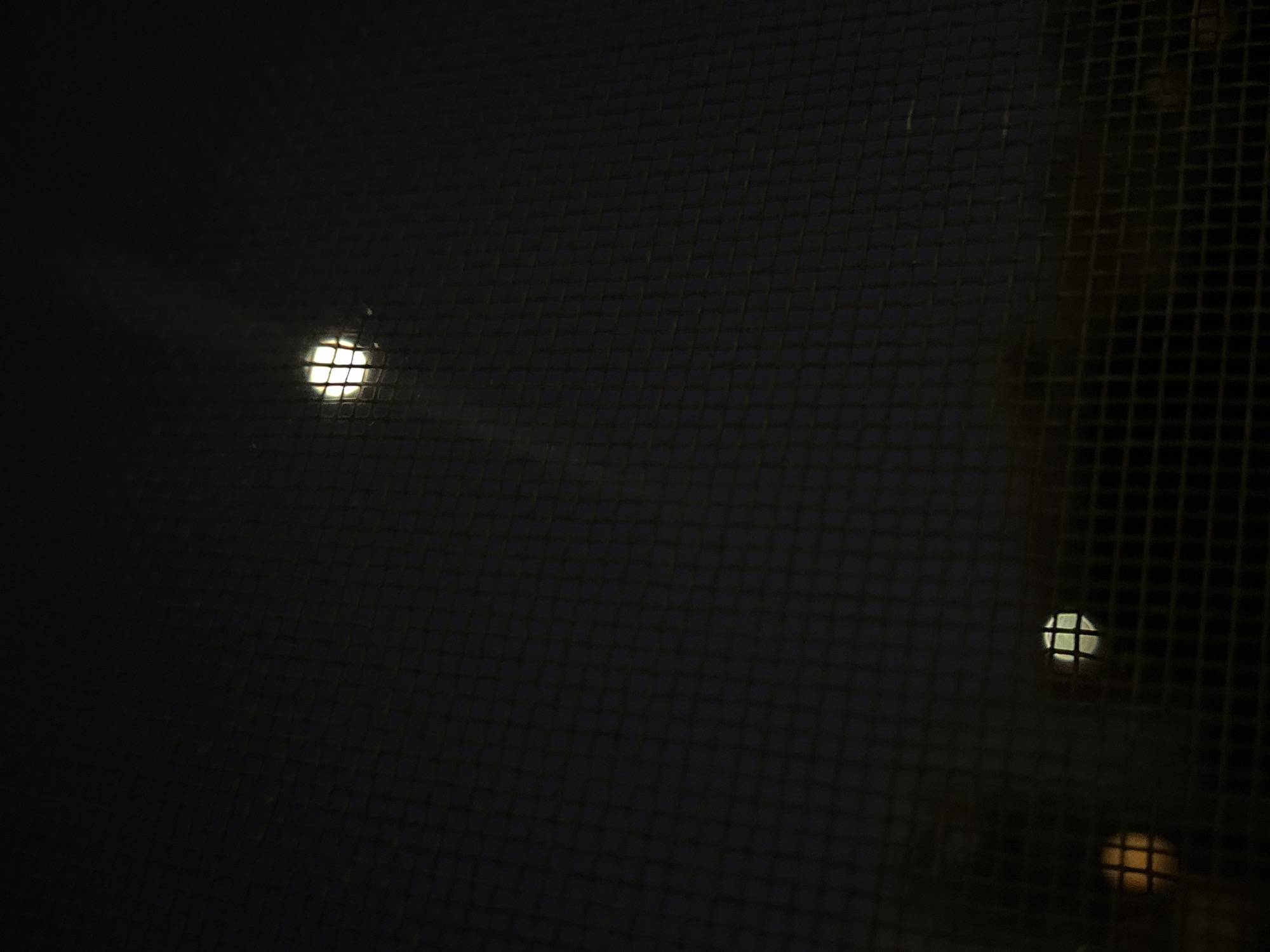 The image shows a dark scene viewed through a mesh screen. Two distinct light sources are visible. One, a bright white point of light, is located towards the upper left. A group of two light sources, one white and one yellow, are visible on the right. The darkness and presence of a screen suggest the view is likely from inside a building looking out, possibly at night. Without further context, it is impossible to confirm the location in Plovdiv, Bulgaria.