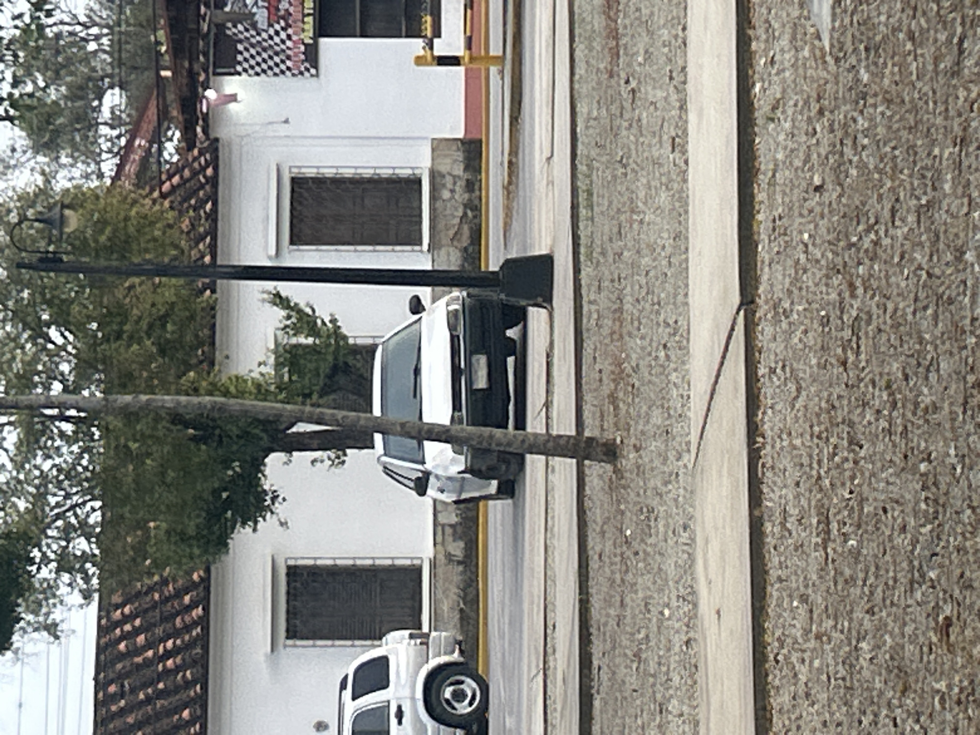 The image shows a street scene in the city of Santa Ana, Venezuela. In the foreground, a paved area is covered with gravel. A concrete pathway runs parallel to the street. In the middle ground, a white car is parked on the street. The car is facing the viewer. A light pole stands to the right of the car. Behind the car, a white building with a tiled roof is visible. The building has several windows with dark shutters. A checkered flag sign is mounted on the right side of the building. In the background, trees and power lines can be seen. A silver SUV is parked to the left of the white car, partially visible.
