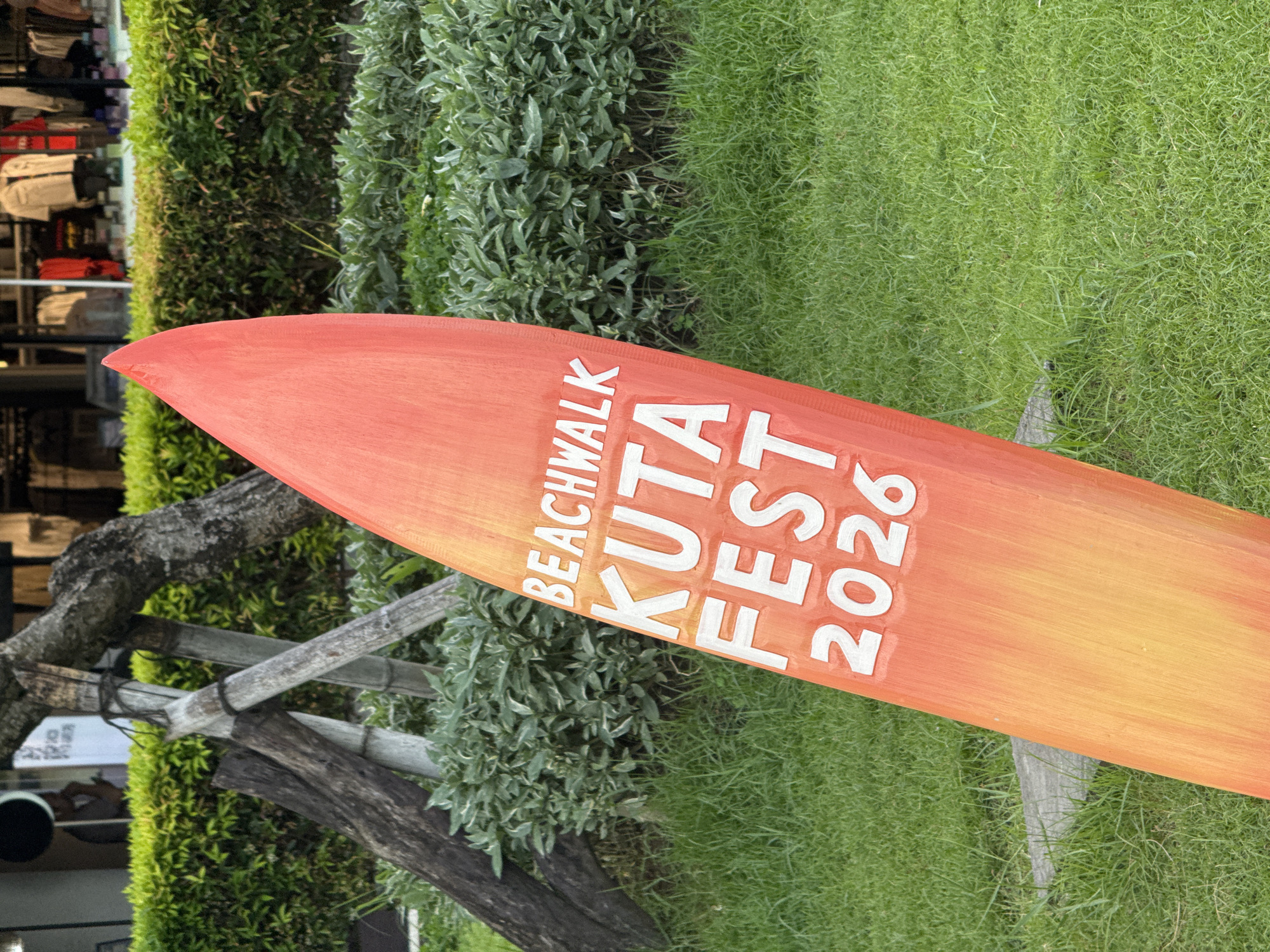 A surfboard-shaped sign reads "Beachwalk Kuta Fest 2026" and is placed outside on green grass near a bush. A tree and what appears to be part of a building are behind the sign. The setting is Kuta, Indonesia.