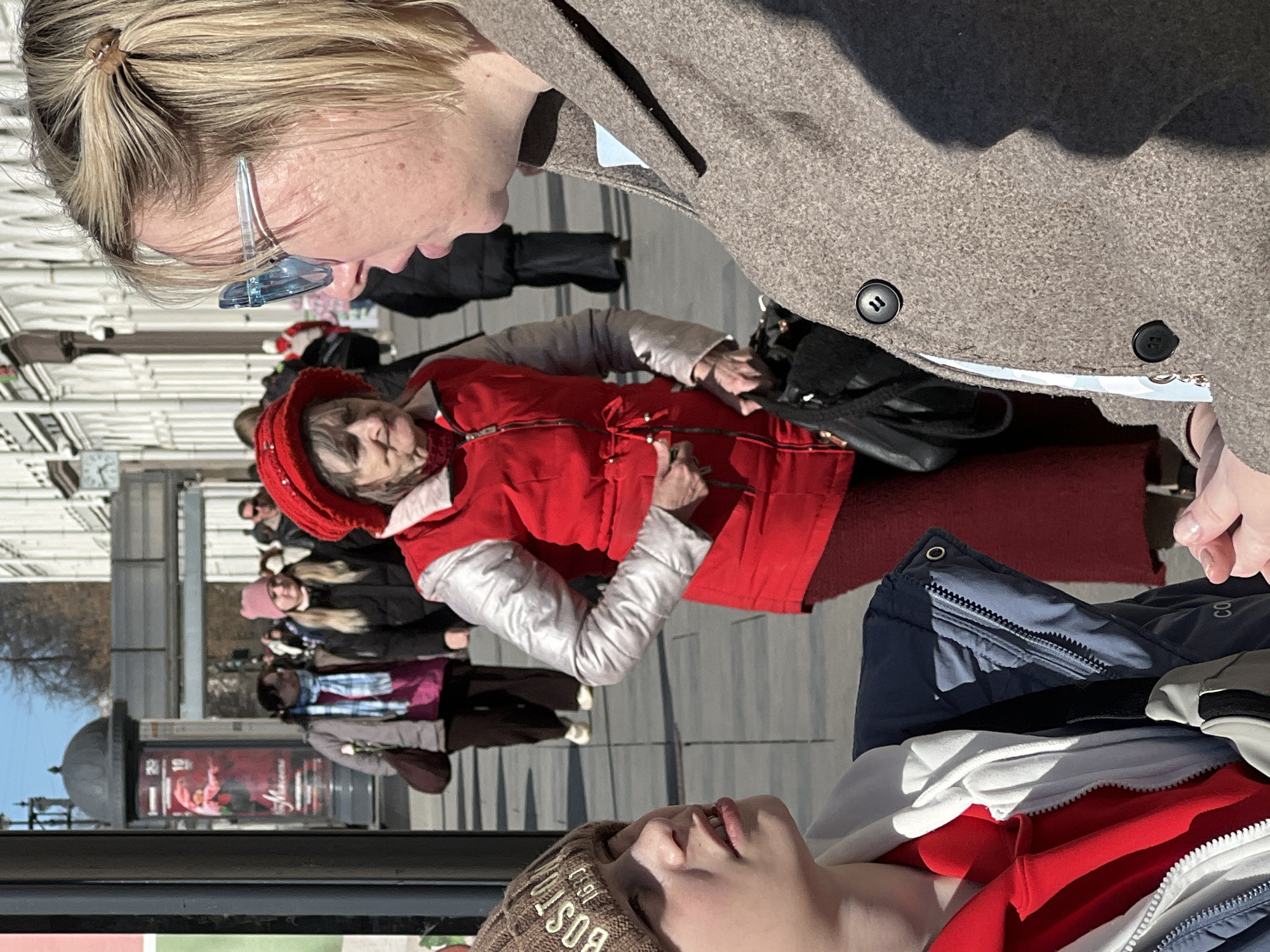 This image, taken in Saint Petersburg, Russia, captures an outdoor urban scene on a bright, sunny day. The strong sunlight casts clear shadows, suggesting midday or early afternoon.
In the foreground, a young person is visible, wearing a brown knitted beanie with the text "BOS" or "BOST" and a navy blue jacket with "CO" and a logo on it, possibly Columbia. Their head is tilted upwards.
In the midground, an older woman is prominent, dressed in a vibrant red puffer vest over a white long-sleeved top, a matching red hat, and a red long skirt. She carries a black handbag and looks towards the left with a thoughtful or serious expression.
Above her, a blonde-haired woman with light blue-tinted sunglasses, her hair tied back, is seen in profile. She wears a textured brown coat and looks downwards, seemingly at the woman in red or the young person.
In the background, several blurred figures walk along a paved street or pedestrian area. A dark, cylindrical street poster stand is visible, featuring a red advertisement. The poster displays "18+" and partially legible text that appears to be related to a cultural or theatrical event, possibly "Mannes" or "Marinette." The setting is an active city street with paved ground and reflective surfaces, possibly shop windows, reflecting the sky and distant buildings.