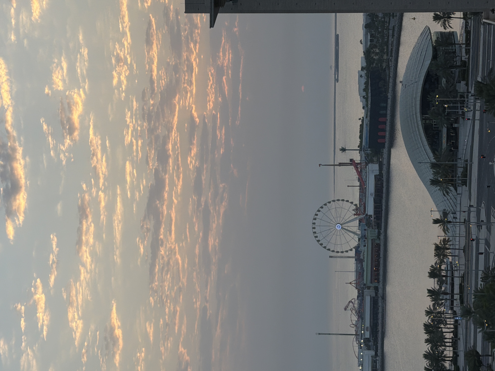 The image captures a scenic view of Lusail, Qatar, at dusk. The sky is filled with scattered clouds, tinged with warm hues of orange and pink, indicating the setting sun. In the middle ground, a Ferris wheel illuminated with white lights stands prominently against the calmer waters of the Arabian Gulf. Adjacent to it, a roller coaster and other amusement park structures are visible, suggesting a lively entertainment district. The waterfront features buildings with dark facades and some with red accents. Palm trees line the foreground, bordering a modern architectural structure with a curved roof. The overall atmosphere is tranquil, with the city lights beginning to twinkle as daylight fades.