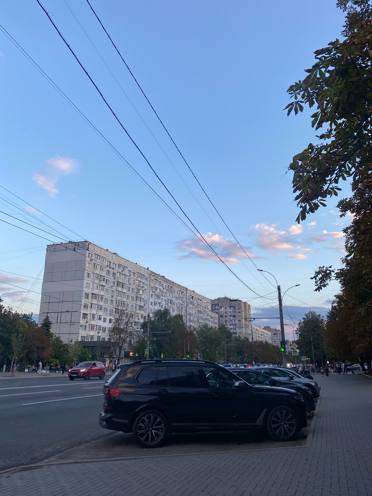 This image depicts a street scene in Chișinău, Moldova, likely in the evening. A long, multi-story apartment building with many windows dominates the background. Several power lines are visible stretching across the sky, some appearing to sag. Trees line the street and are partially visible in the foreground and on the right side of the image. In the foreground, several parked cars are visible, including a black SUV. A red car is driving on the street. Pedestrians are visible on the sidewalk in the distance. The sky is partly cloudy with hues of pink and orange, suggesting a sunset or sunrise. The overall atmosphere is urban and residential.