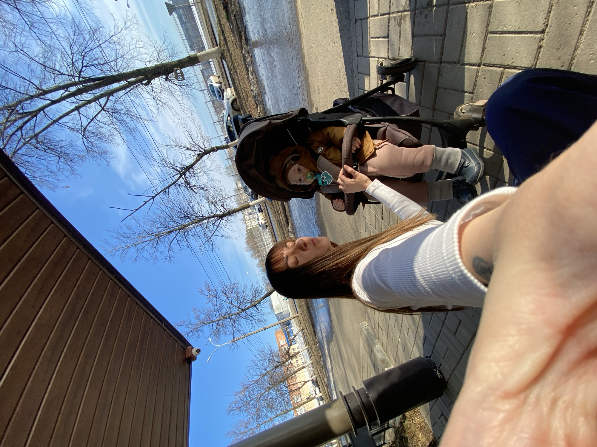 The image shows a woman standing next to a stroller in an urban environment. The stroller contains a baby with a pacifier. The location is likely Votkinsk, Russia, based on the prompt.
The woman has long brown hair and wears a white long-sleeved shirt. Her hand is visible, holding what appears to be a pacifier string. The baby in the stroller wears a brown jacket and pink pants.
The background shows a street with puddles, buildings, bare trees, and utility poles. The sky is blue with some clouds. The ground is paved with bricks. There is a building with brown siding on the left and a metal railing in the foreground. The overall impression is of a sunny day in early spring.