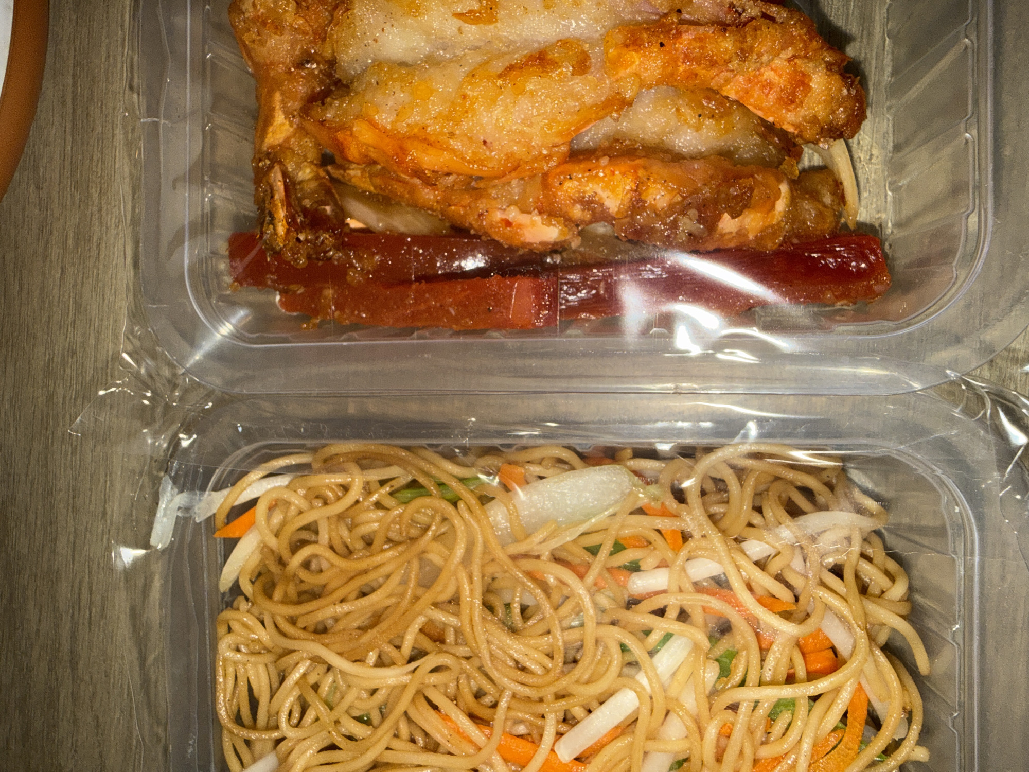 The image shows two clear plastic takeout containers sitting on a wooden surface, likely a table or countertop.
The top container holds what appears to be a dish consisting of breaded or battered shrimp (or similar seafood) and reddish-brown rectangular pieces, possibly ham or another processed meat.
The bottom container holds noodles mixed with julienned vegetables, including carrots, onions, and possibly scallions.
There are no people visible in the image. The containers suggest a meal prepared for takeout or delivery.
The prompt mentions "Domont, France". However, based only on the image, it is impossible to determine the specific location.