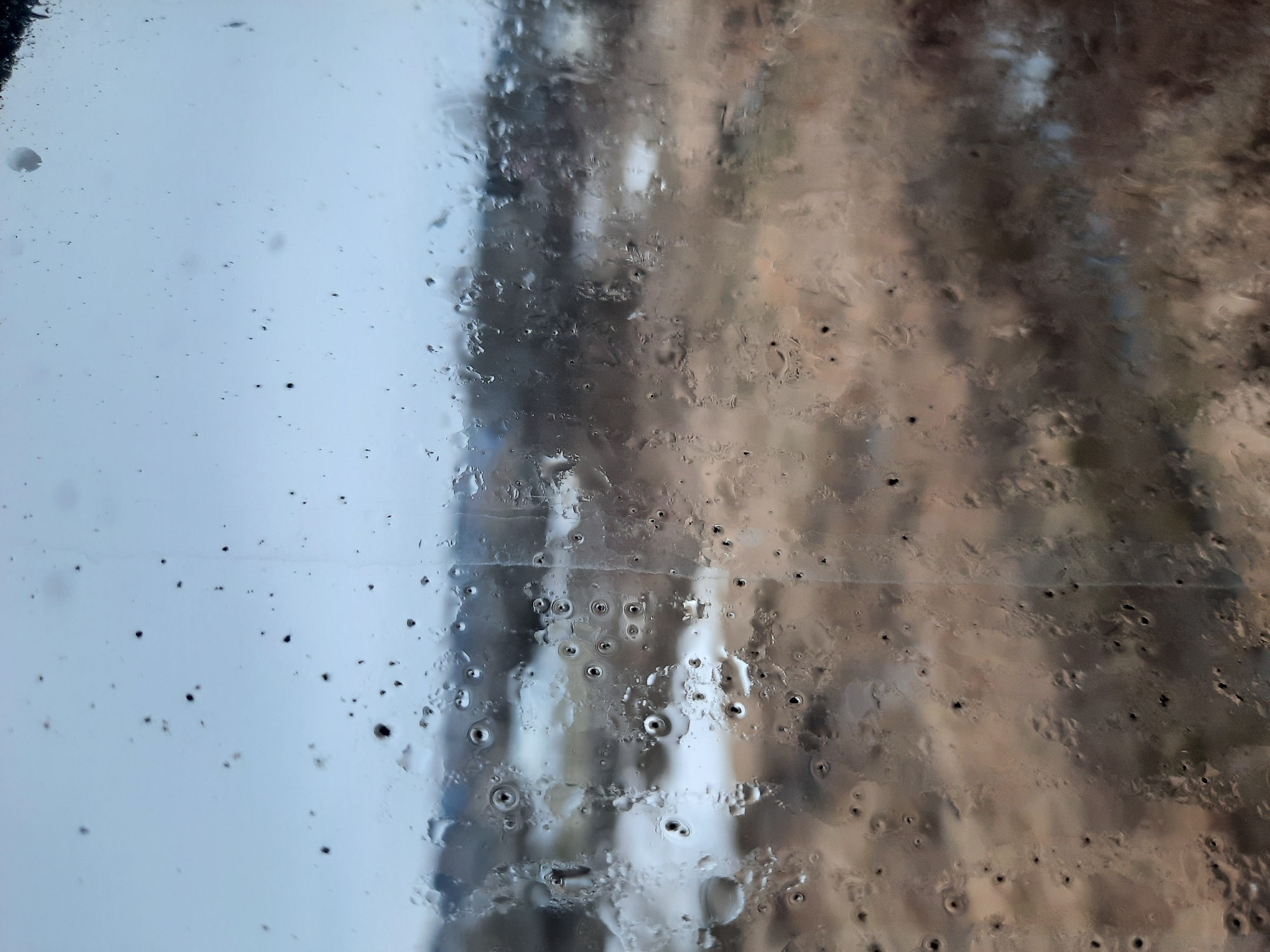 The image depicts a window covered in condensation and droplets, obscuring the view outside. The left side of the window appears cleaner with scattered dark specks, possibly dirt or debris. The right side is heavily covered in water droplets, distorted reflections suggest trees or buildings in the background. The location is Krasnogorskoye, Russia, but due to the obscured view, specific details about the environment are unclear. There are no people or actions visible in the image.