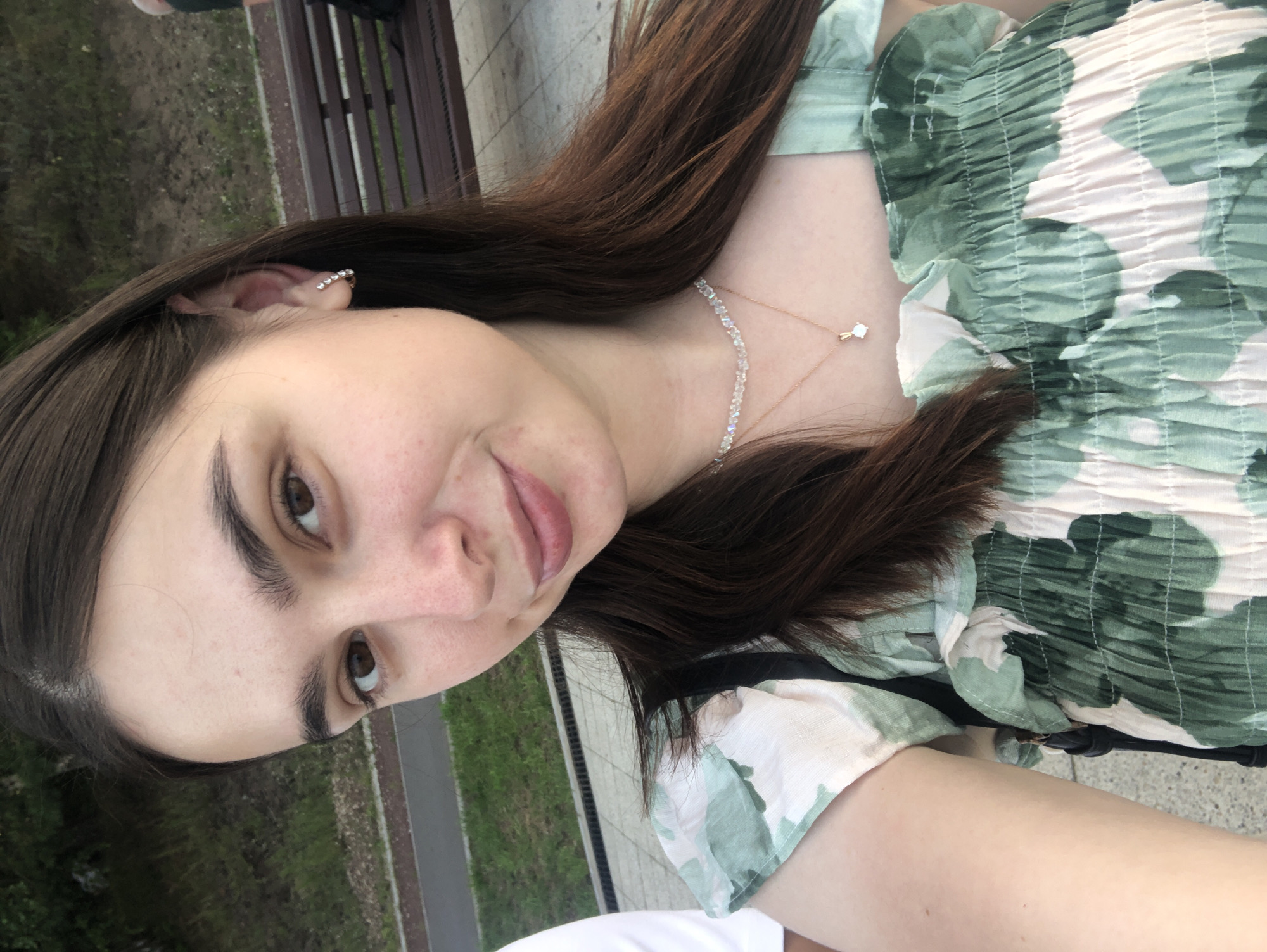 This is a selfie of a young woman outdoors, likely in a park or public space. She has long, dark brown hair and is looking directly at the camera. She is wearing a green and white floral print top with a ruched, elasticated neckline. Around her neck, she wears a delicate gold chain with a small, clear pendant. A sparkling earring is visible on her left ear. Her expression is neutral.
The background shows hints of an outdoor setting with green grass and a paved walkway or path. There is also a glimpse of a wooden bench. The lighting suggests it is daytime. There is no visible text in the image. The overall mood is casual and relaxed. The context places this image in Chaikovsky, Russia.