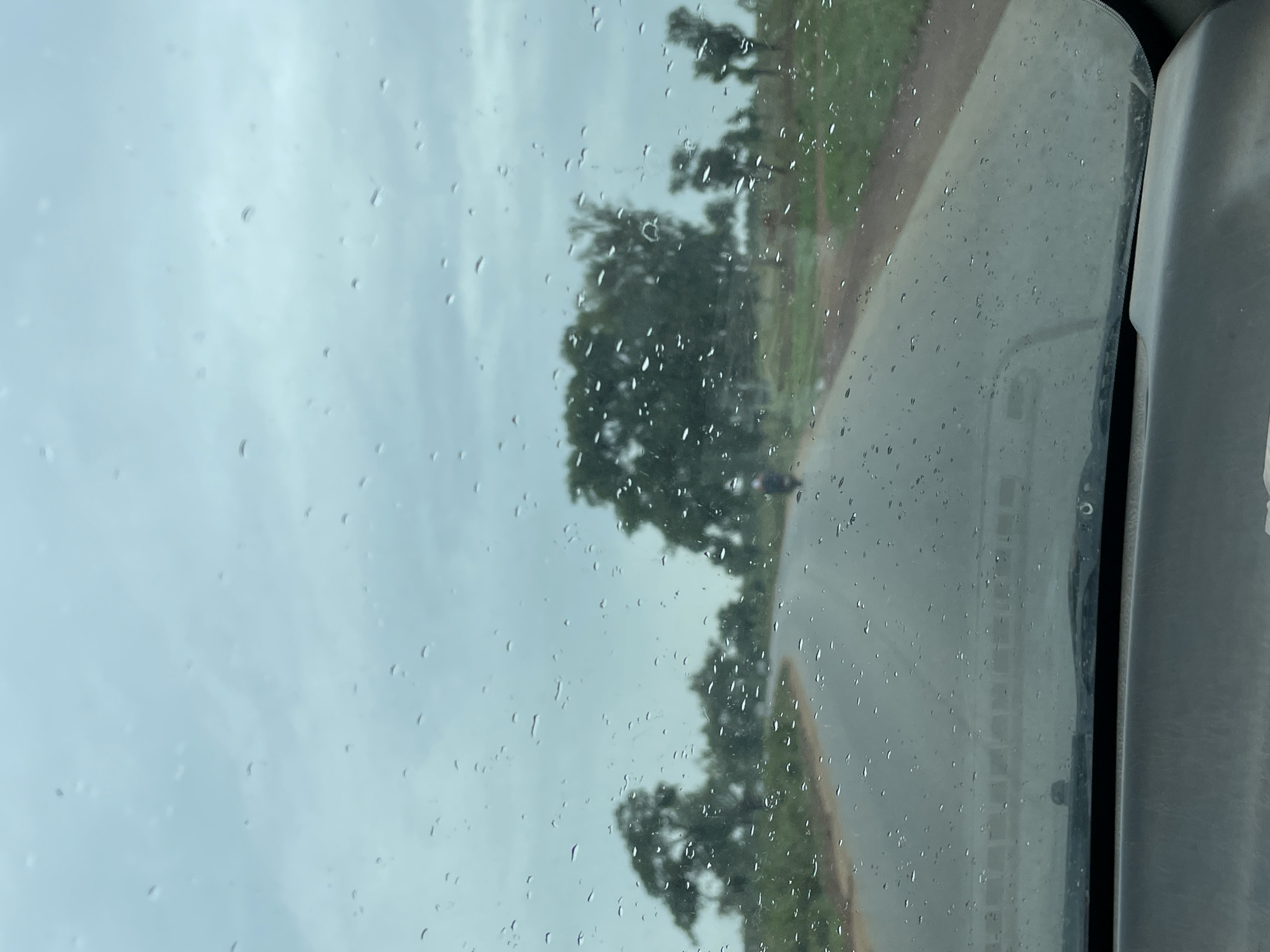 The image, taken from inside a vehicle, shows a road running alongside trees in Kujama, Nigeria. Raindrops are visible on the car window. A person can be seen riding a motorcycle on the road. The sky is overcast.
