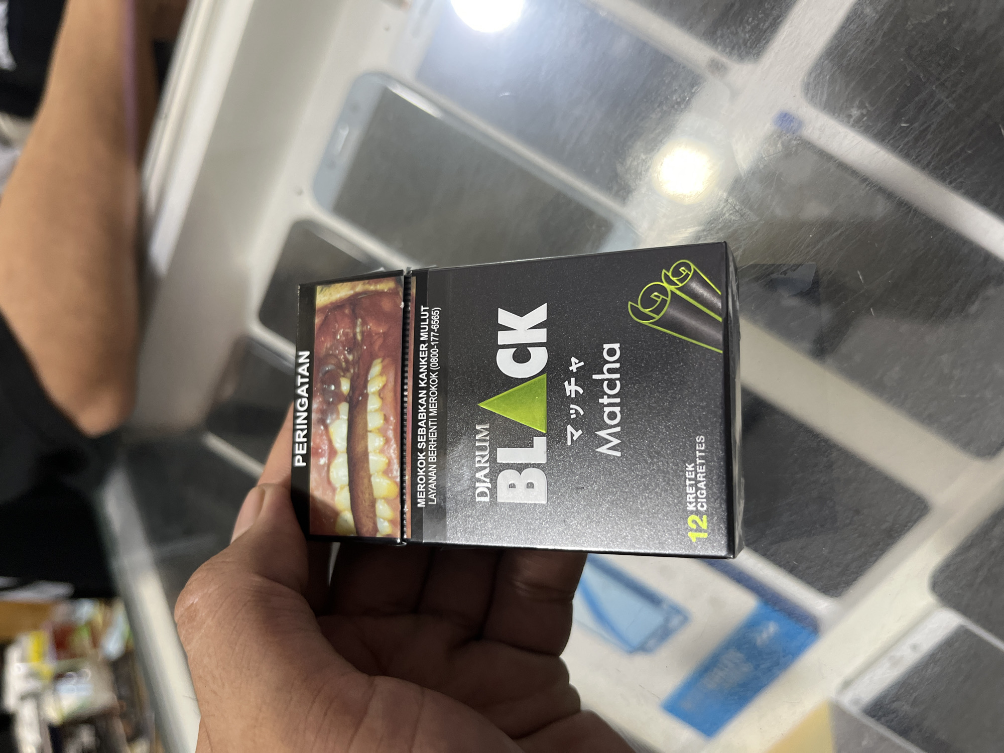 A close-up, eye-level shot shows a person's hand holding a black pack of Djarum Black Matcha kretek cigarettes. The pack displays a warning image of unhealthy teeth and gums, the brand name "DJARUM BLACK" in silver and green, and "Matcha" in Japanese and English. Below that, "12 KRETEK CIGARETTES" is visible. The background is blurred and appears to be a store counter with various items, including what seem to be cell phones and other merchandise, suggesting a retail environment. The lighting is adequate, highlighting the product. The scene is likely within Central Jakarta, Indonesia, given the brand of cigarettes.
