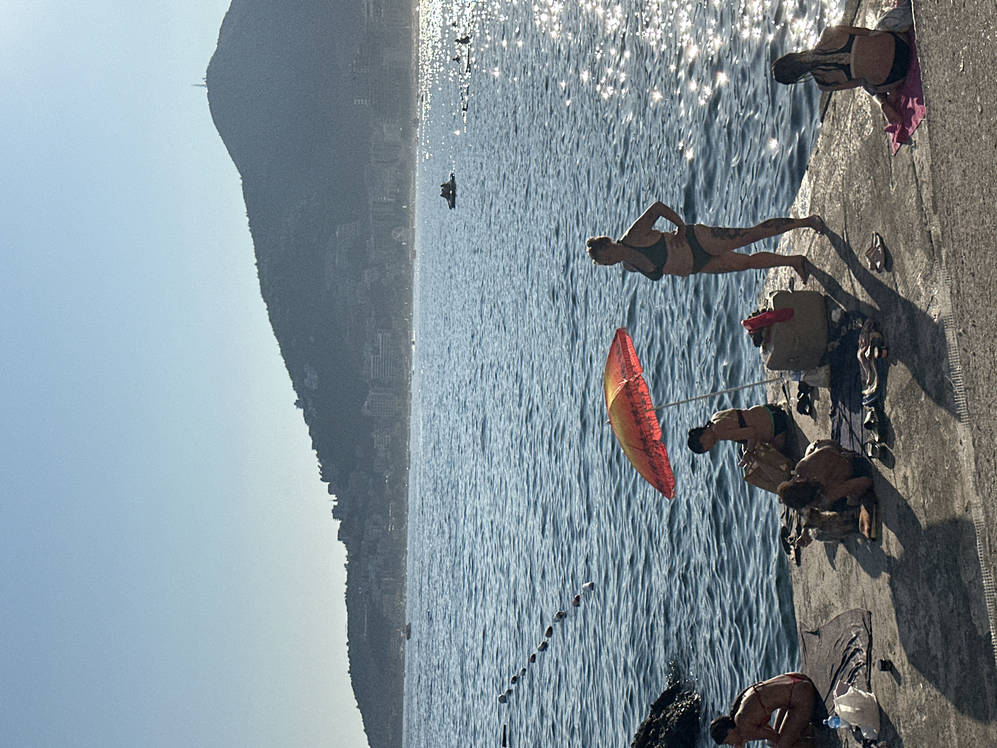 The image captures a sun-drenched day at a coastal area in Budva, Montenegro. In the foreground, several individuals are visible on a concrete platform adjacent to the water. A woman in a green bikini stands with her hands on her hips, facing away from the camera. Nearby, two people are seated on the platform, one appearing to be looking at a book or papers, while another is leaning back. Another person in a red bikini is seated on a towel to the far left. A red and yellow umbrella provides shade for a group of people sitting under it. The sea is calm and shimmering with sunlight. In the background, a mountainous landscape with buildings and a prominent tower on its summit forms the backdrop. Several small boats and people on paddleboards are scattered across the water. A rope with buoys demarcates a section of the sea. The scene suggests a leisurely day at the beach, with people enjoying the sun and sea.