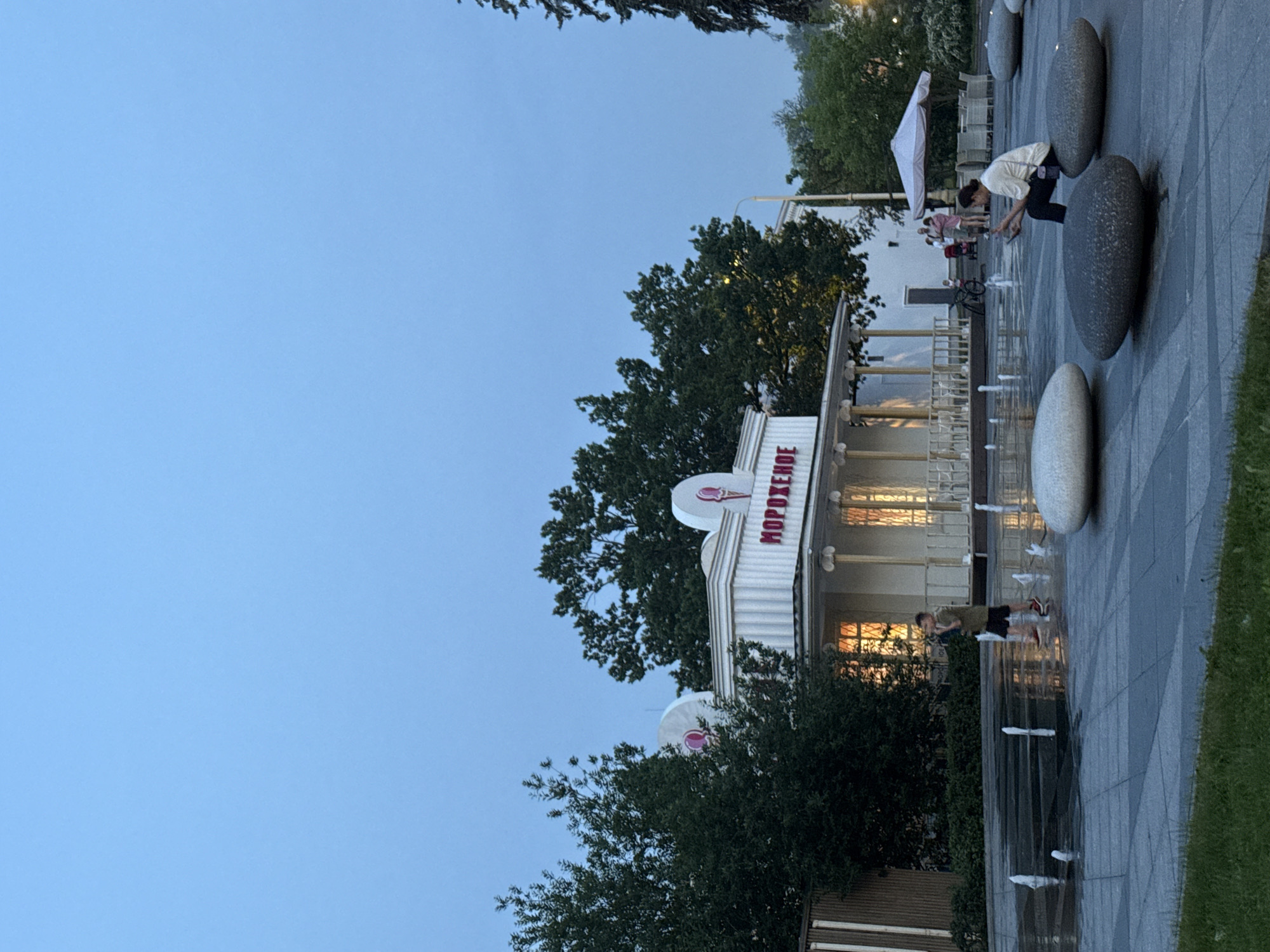 The image captures an outdoor scene at dusk, likely in Moscow, Russia, given the Cyrillic text "МОРОЖЕНОЕ" (which translates to "ice cream") visible on a building. In the foreground, a paved area features a water fountain with several jets of water spraying upwards. Large, smooth, oval-shaped stones are scattered across the paved area, serving as seating.
Two people are visible. A young boy in a green shirt and shorts is walking through the fountain area, while a woman in a cream-colored top and dark pants is seated on one of the stones, looking at her phone. In the background, an ice cream parlor with a white facade and pink ice cream cone motifs is illuminated. Behind the parlor, trees create a backdrop, and a white umbrella shades outdoor seating. The sky is a clear, pale blue, indicating the transition from day to night.