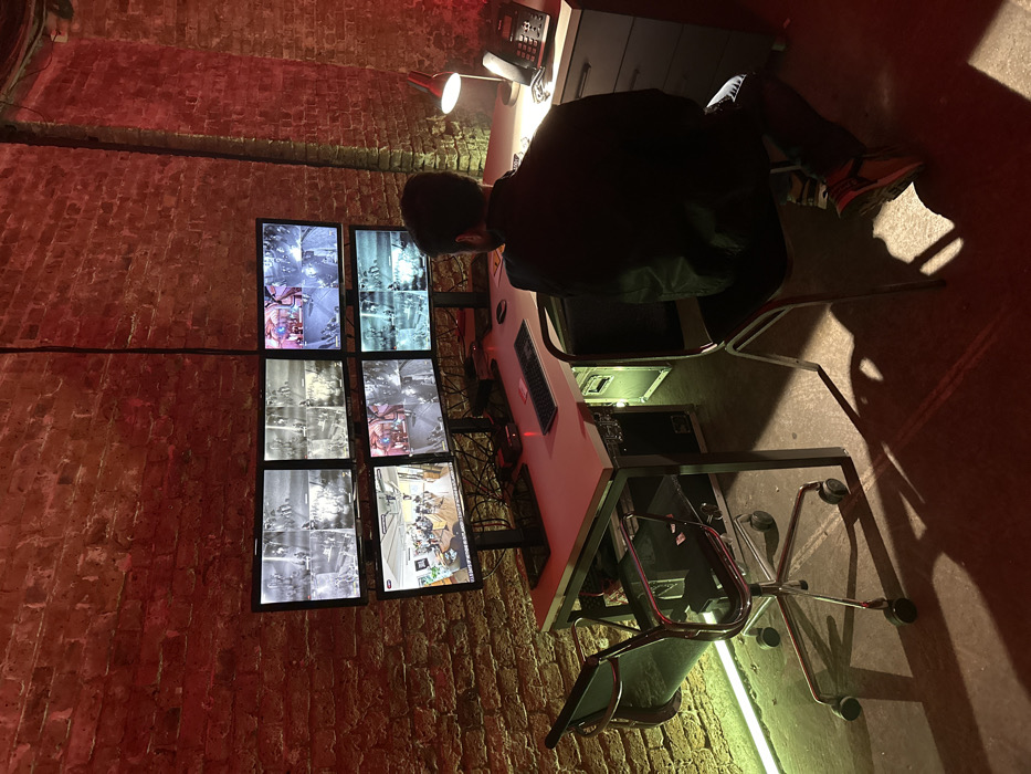 A single adult male, viewed from behind, is seated in a control room in an exposed brick structure, likely a repurposed industrial space or basement in London, United Kingdom. The room is dimly lit with strong red and green accent lighting illuminating the brick walls and concrete floor. The male, with short dark hair, wears a dark jacket, dark trousers, and sneakers with red accents. He is positioned at a light-colored, L-shaped desk facing a bank of six flat-screen monitors arranged in a 3x2 grid. The monitors display various surveillance feeds: some monochrome, others in color, depicting different indoor and outdoor scenes, including a room with multiple individuals and musical instruments. Keyboards and mice are present on the desk. A flight case beneath the desk emits a green light. To the right of the desk, a red adjustable desk lamp illuminates a black desk telephone. The individual is actively engaged in monitoring the visual displays.