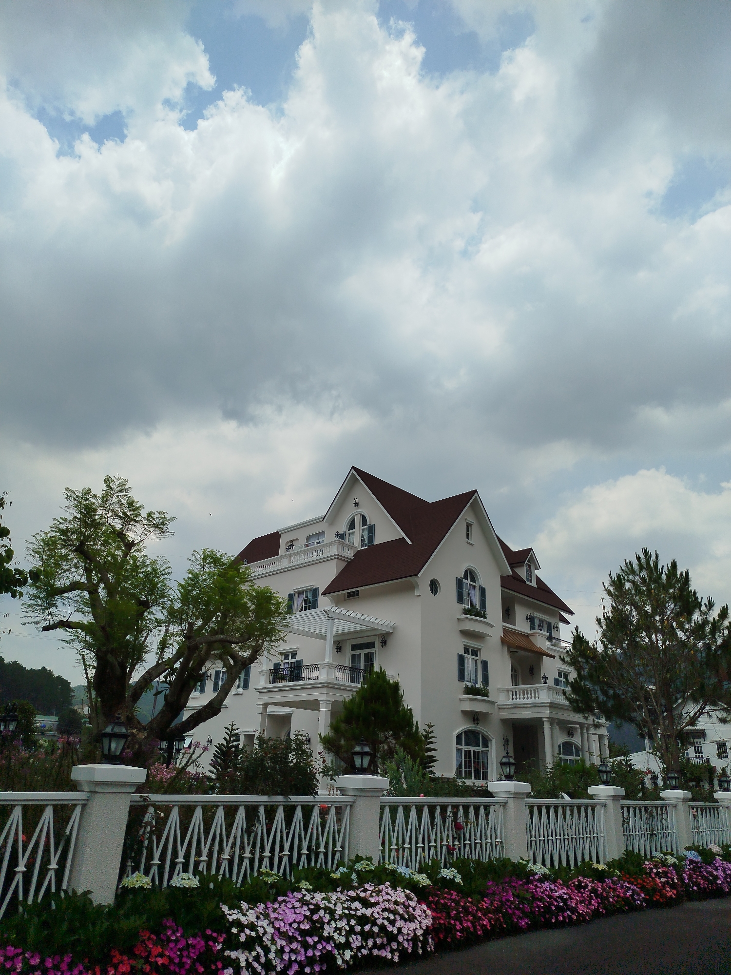 A large, multi-story white house with a brown roof stands in Hiep Thanh, Vietnam. It has multiple windows with blue shutters, balconies with white railings, and is surrounded by greenery. A white fence with ornate details runs along the front, separating the house from a flower bed filled with pink, purple, and white flowers. Trees flank the house on either side. The sky is mostly cloudy.