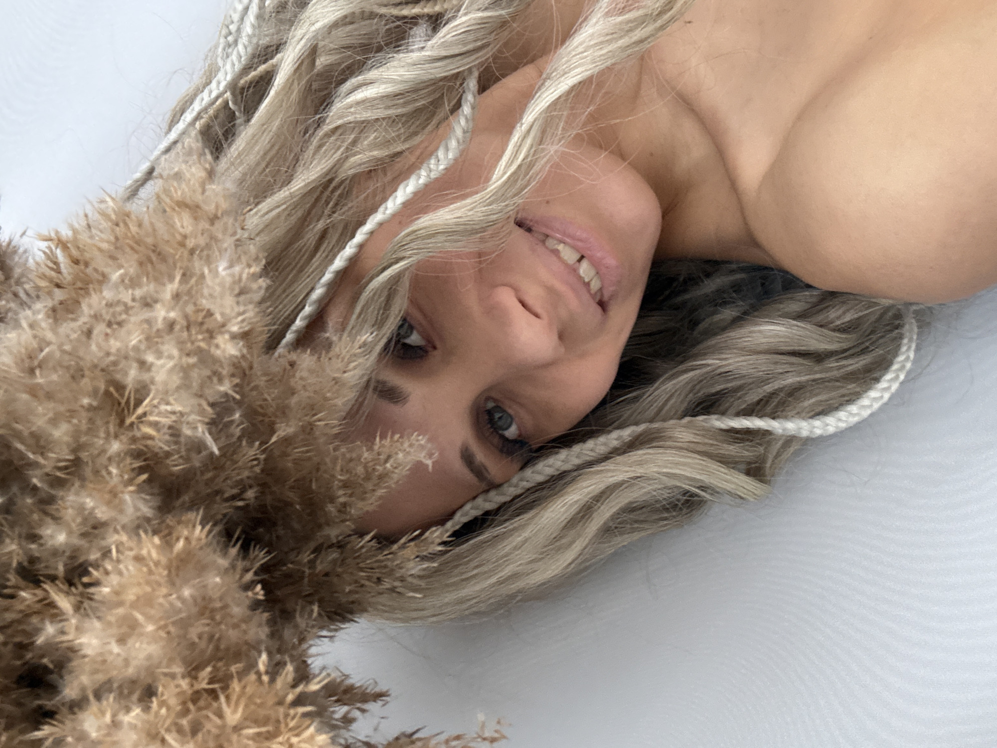 A woman with fair skin, light blonde hair with white braids, and blue eyes lies on a white surface. She is partially obscured by a cluster of dry, beige-colored pampas grass. The photograph appears to be taken at a close, overhead angle. The location is not discernible in the image.