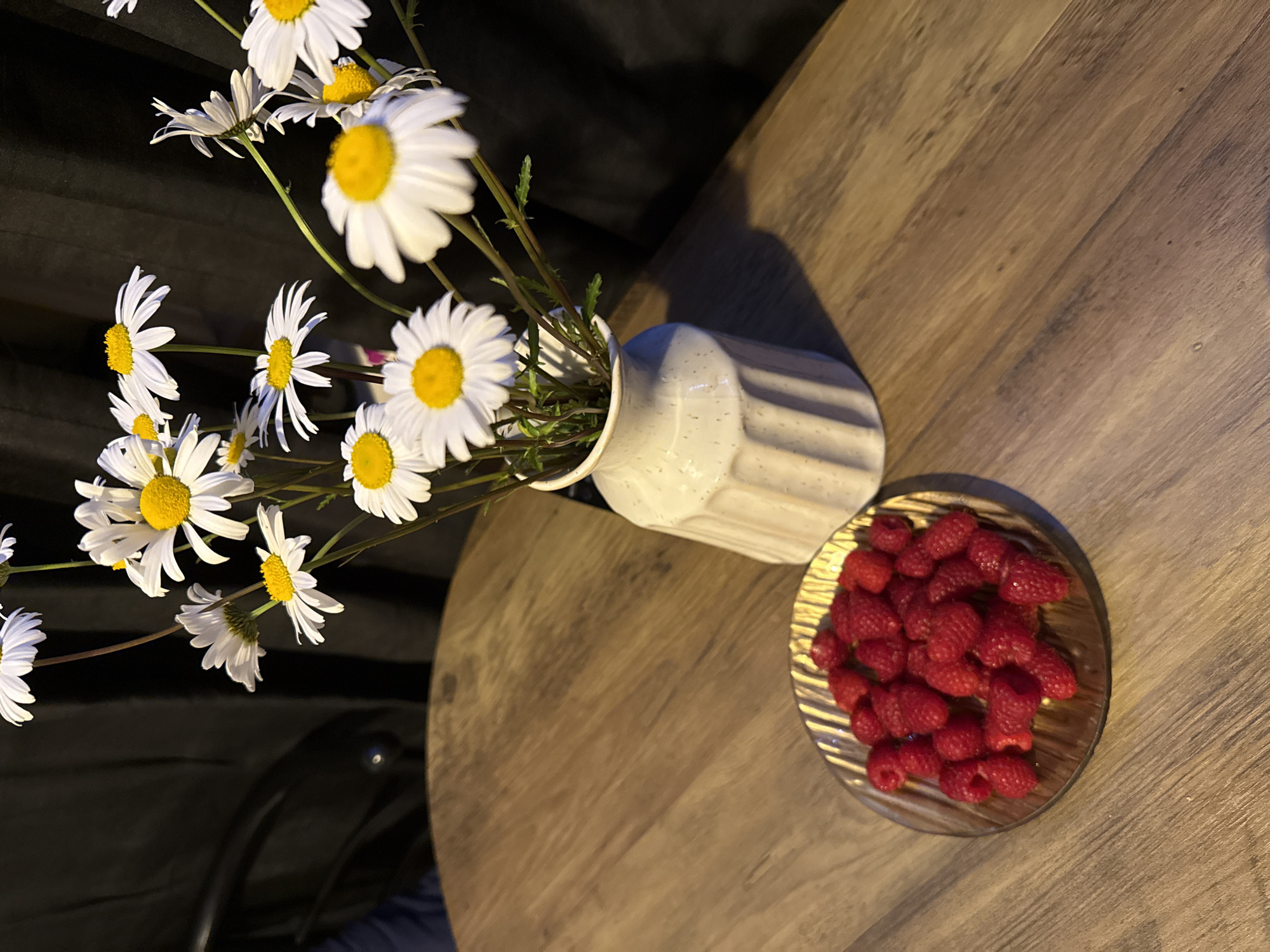 A still-life photograph shows a vase of daisies and a plate of raspberries on a wooden table. The daisies are in a white, textured vase that is lying on its side. The raspberries are on a clear, decorative plate. The background is dark, suggesting an indoor setting. The scene appears to be staged. There is no evidence to indicate that the location of the photograph is Great Yarmouth, United Kingdom. There are no people present in the photograph.
