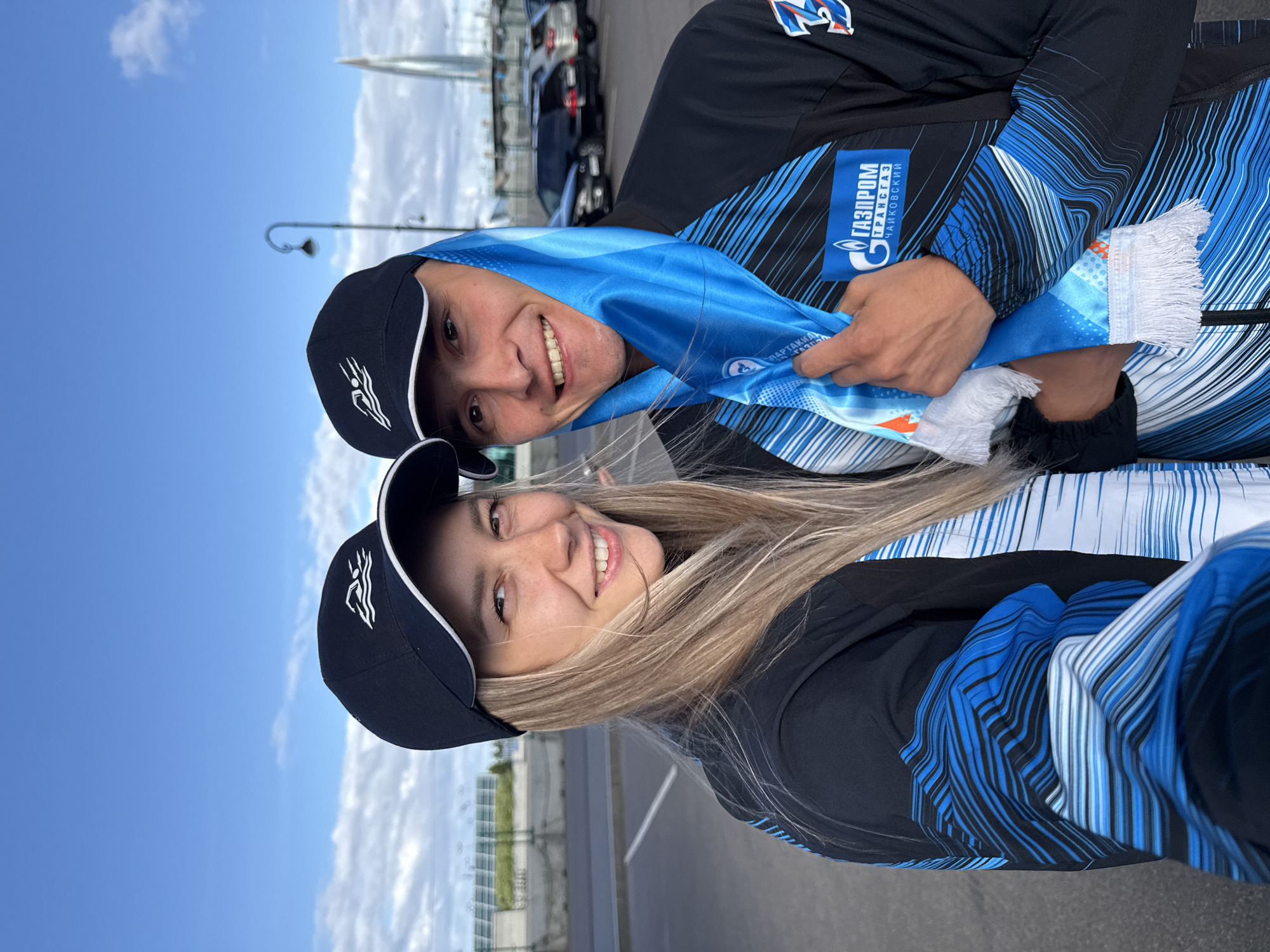 A young couple, a man and a woman, pose for a selfie outdoors in what appears to be a parking lot with a distinctive modern skyscraper in the background. Both are wearing matching black baseball caps with a white logo depicting a stylized wave or swimmer. The woman, on the left, has long blonde hair and is smiling at the camera. The man, on the right, is also smiling and is wearing a blue and white scarf draped around his neck and shoulders. He is holding the scarf with his right hand, revealing a logo that says "ГАЗПРОМ ТРАНСГАЗ ЧАЙКОВСКИЙ" in Russian. Both are wearing black athletic jackets with blue and white patterned accents. The background shows a cloudy sky, some cars, and a street lamp. The architectural style of the skyscraper suggests it could be the Lakhta Center in Saint Petersburg, Russia, known for its distinctive shape and the Gazprom logo. The overall impression is a casual, positive moment captured by the couple, possibly at an event related to the sponsorship displayed on the scarf.