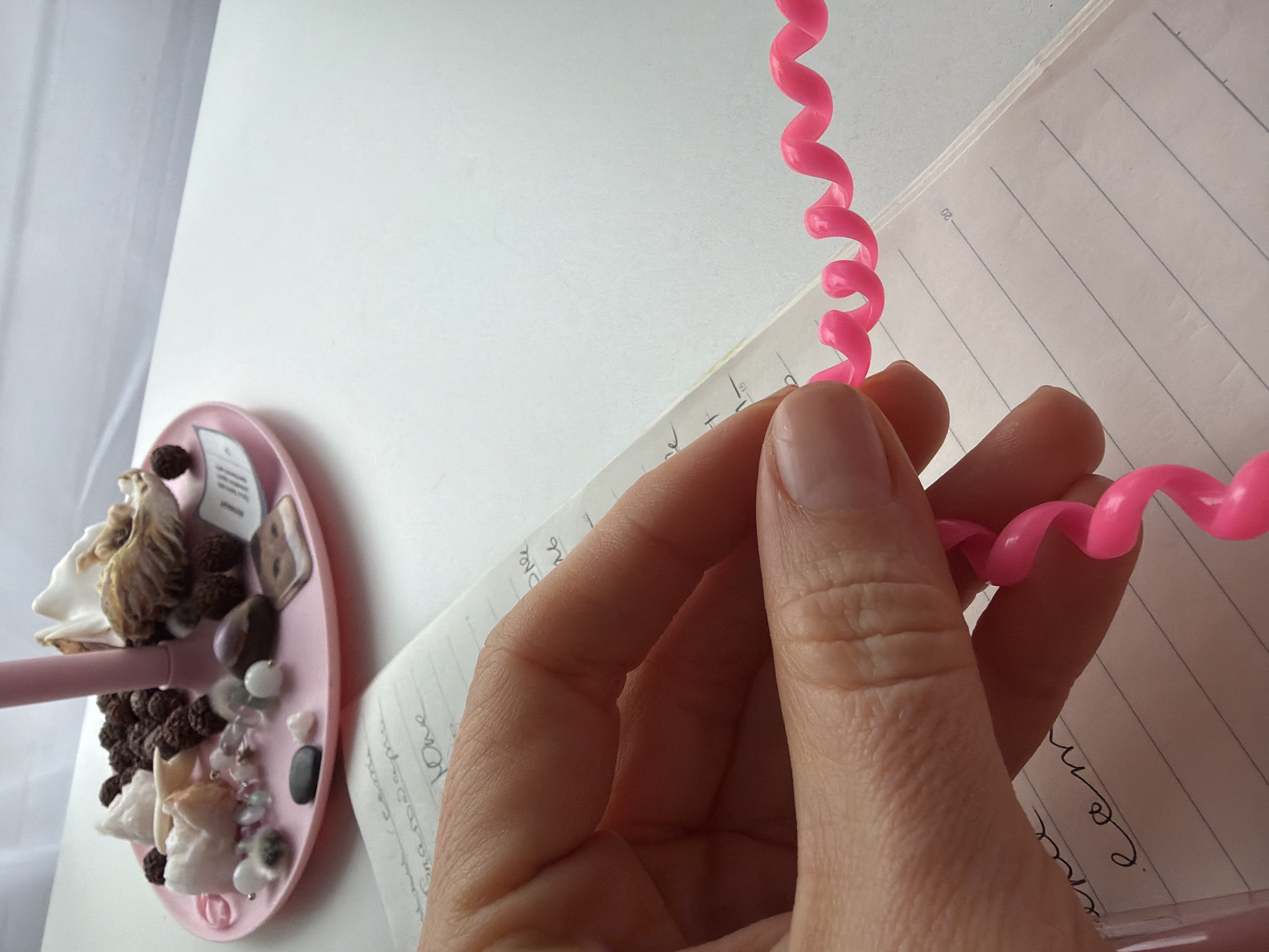 A close-up shot features a hand holding a pink, coiled telephone cord. The hand is pale, with visible skin texture and wrinkles. The background includes an open notebook with lined pages and handwritten text, and a pink tray holding a collection of small objects like shells, beads, and stones. The environment suggests an indoor setting, possibly a workspace or desk area. The image was likely taken in the city of Saint Petersburg, Russia.