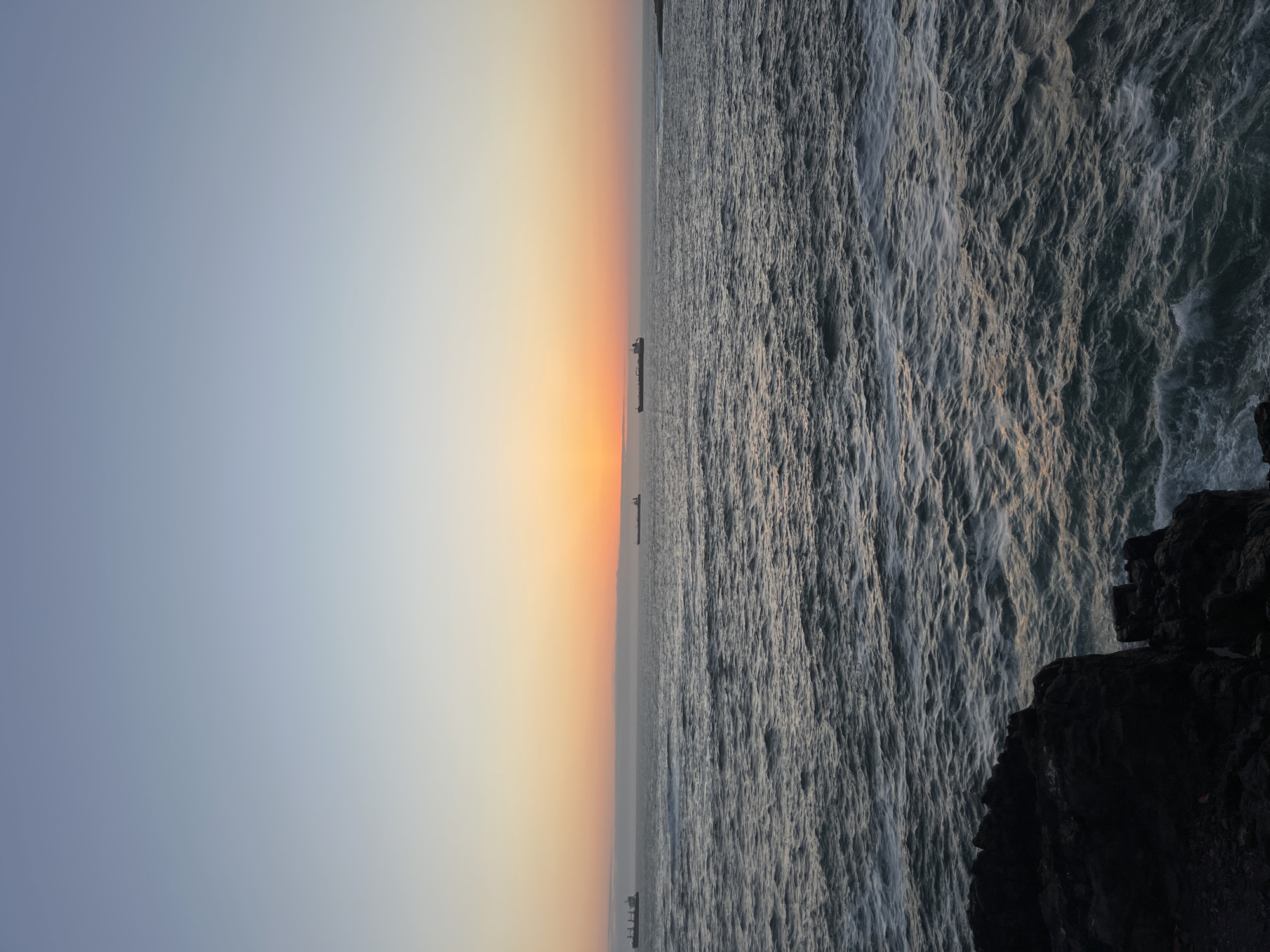 The image depicts a sunset over the Pacific Ocean, viewed from the coast of Arica, Chile. The sky transitions from light blue to orange and yellow near the horizon. Three ships are visible on the horizon. The water is choppy, with a dark gray coloration. A dark rock formation is visible in the lower right corner. The image is vertically oriented.