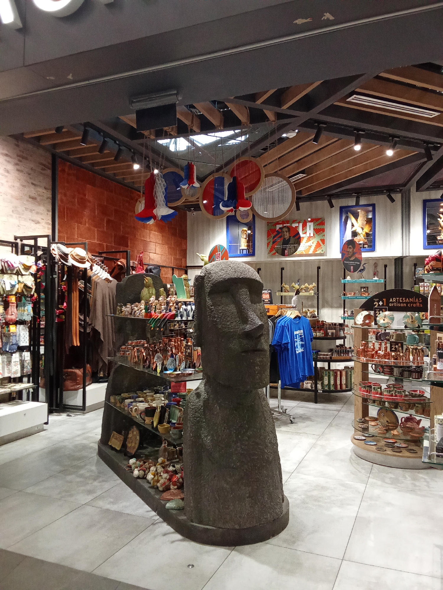 The image depicts the interior of a souvenir shop, likely located in Pudahuel, Chile, given the context of the Easter Island statue. A large, dark grey, stone-like statue resembling a Moai, a traditional Easter Island statue, stands prominently in the foreground, occupying the lower-left quadrant of the image. Behind the statue, a variety of merchandise is displayed on shelves and racks.

To the left of the Moai, a black metal rack displays clothing items, including hats, scarves, and what appear to be bags. Further to the left, a white display stand holds more folded items. The background wall to the left is constructed of rough, reddish-brown bricks.

To the right of the Moai, shelves are filled with various souvenirs. These include ceramic mugs, plates, small figurines of animals and people, and other decorative items. Some shelves are made of wood, while others are glass. A prominent black sign with white text reads "ARTESANIAS artisan crafts". A bright blue t-shirt is hanging on a rack to the right of the Moai.

Above the merchandise, the ceiling features exposed wooden beams and industrial lighting fixtures. Decorative circular hangings with red, white, and blue elements are suspended from the ceiling in the upper portion of the image. Artwork featuring portraits and other graphic designs are mounted on the walls in the background. The floor is tiled with large, light grey squares. There are no people visible in the image. The overall impression is a well-stocked retail environment offering a variety of cultural and artisanal products.