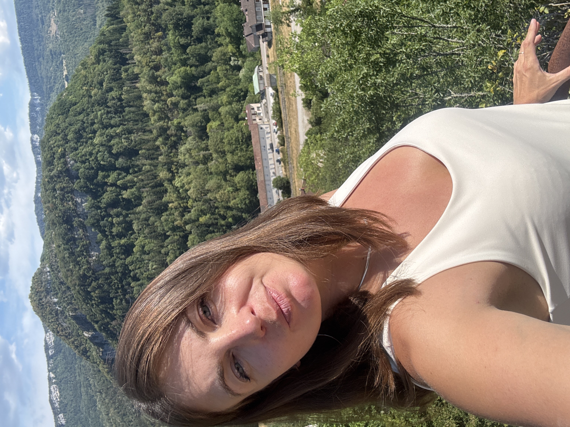 A woman with brown hair is taking a selfie outdoors, with a scenic mountain and forest landscape in the background. She is wearing a white tank top and a silver necklace. In the background, there are buildings nestled within a lush green valley, suggesting a rural or small town setting. The sky is blue with some scattered clouds. The location is identified as Lavans-lès-Saint-Claude, France.