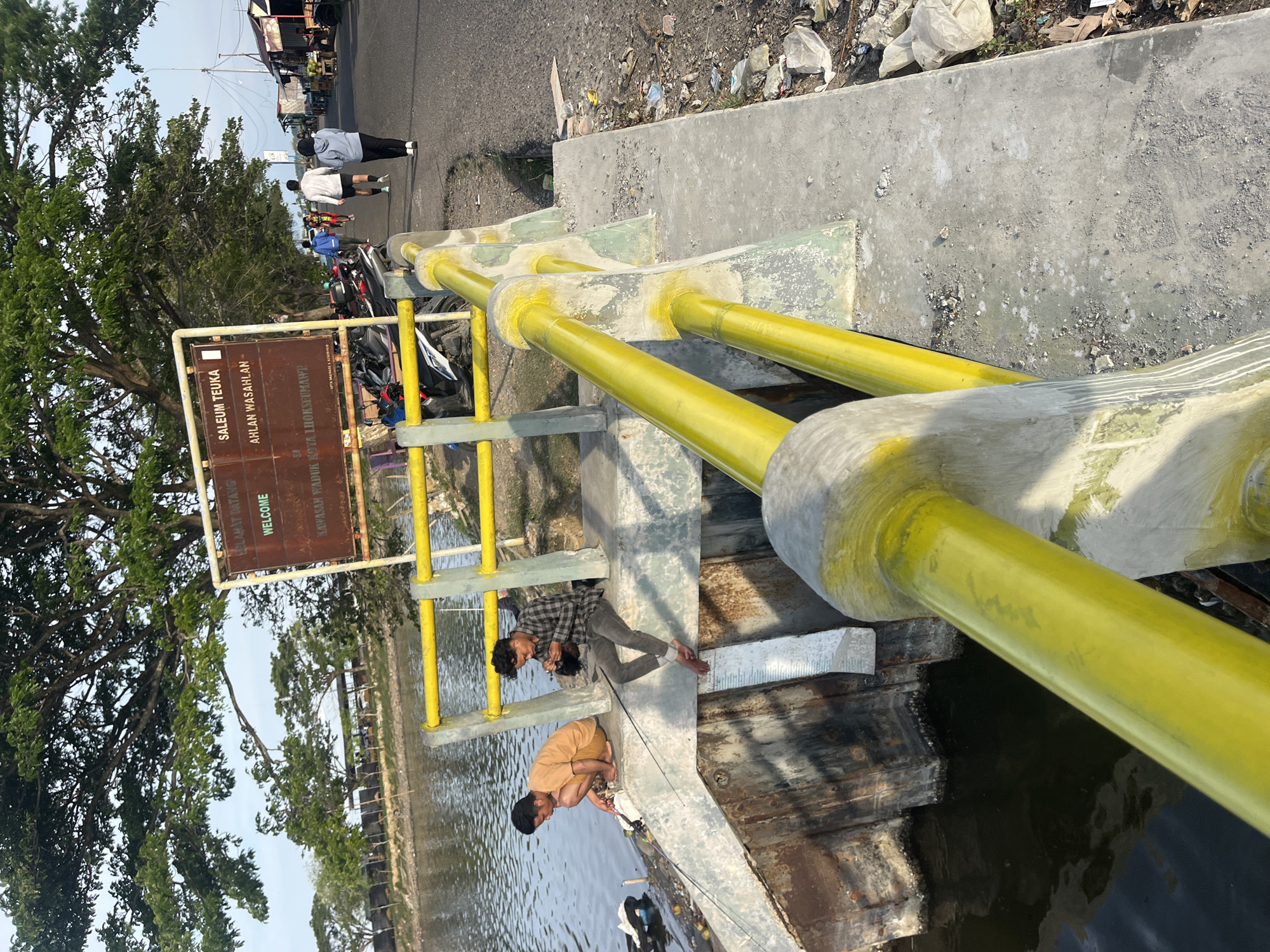 The image shows a bridge over a body of water in Lhokseumawe, Indonesia. Two men are fishing off the side of the bridge. One man is wearing a brown shirt and pants. The other man is wearing a plaid shirt and grey pants. A yellow railing lines the bridge. A sign is visible that says "SALEUM TEUKA, AHLAN WASAHLAN" and "SELAMAT DATANG, WELCOME". Debris is visible on the side of the bridge. In the background, people walk along a road next to motorcycles and buildings. Trees are visible in the distance.