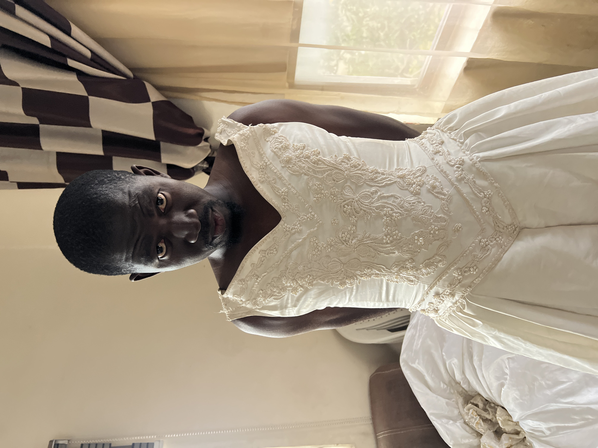 A man is standing in a room wearing a white wedding dress. He is looking directly at the camera with a serious expression. The dress is sleeveless with a V-neckline and is adorned with intricate beadwork and lace. The skirt of the dress is full and made of satin. In the background, there is a window with sheer curtains and a checkered curtain. To the left, there is a brown leather chair and a pile of white fabric, possibly bedding. The room appears to be a bedroom. The lighting is soft and natural, coming from the window. There are no specific landmarks visible that would indicate the city of Jos, Nigeria.