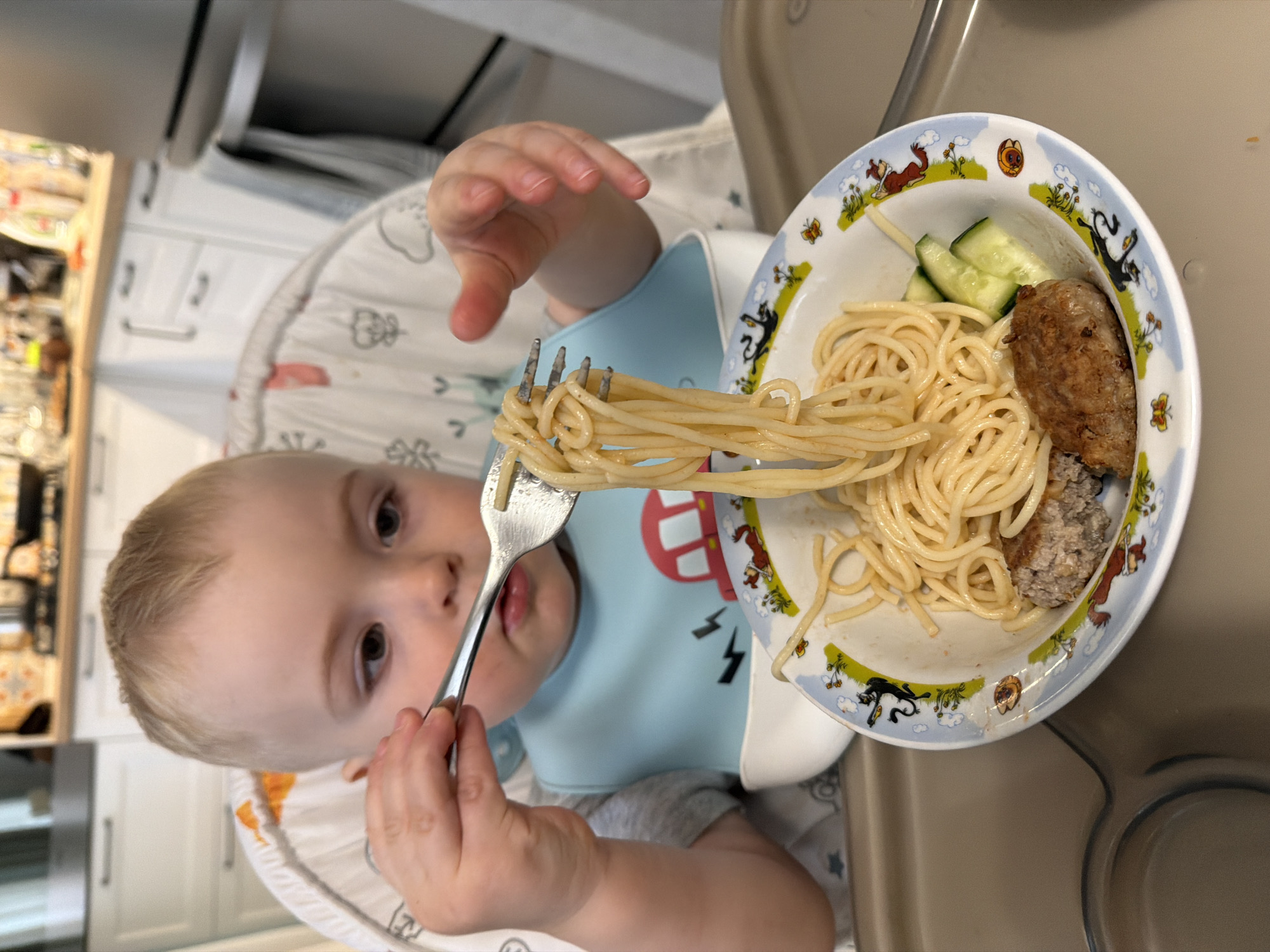 A baby is sitting in a highchair, holding a fork with spaghetti on it. A plate on the highchair tray contains spaghetti, sliced cucumbers, and meatballs. The room appears to be a kitchen. The location of the scene is not discernable from the visual data available.