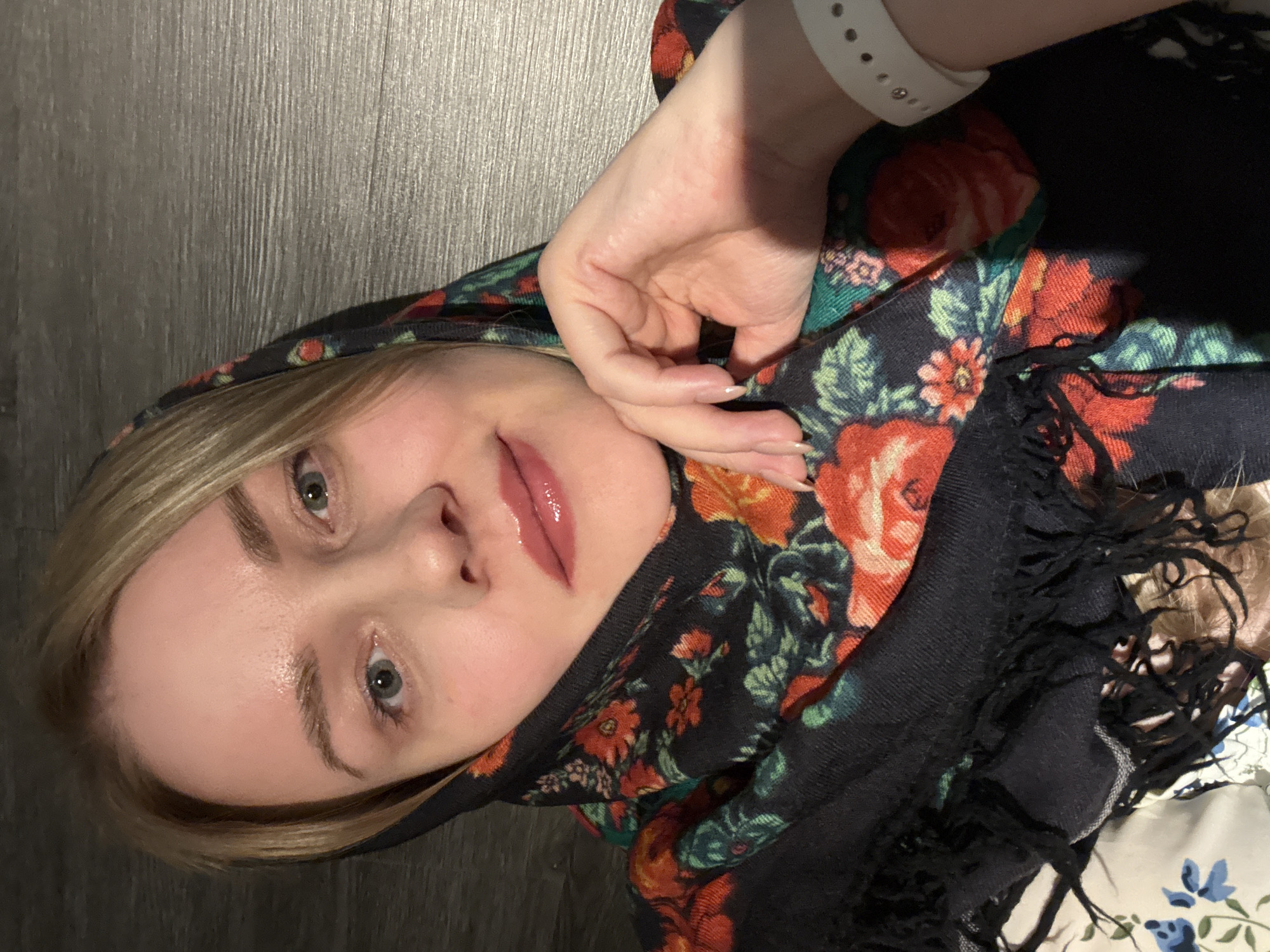 A woman with blonde hair and light skin is wearing a floral shawl, likely a Pavlovo Posad shawl, with red and orange roses, green leaves, and a black background with fringed edges. The shawl is framing her face. She's wearing a white smartwatch. The background appears to be a dark, wooden-like surface. There's no indication of Chaikovsky, Russia, in the image.