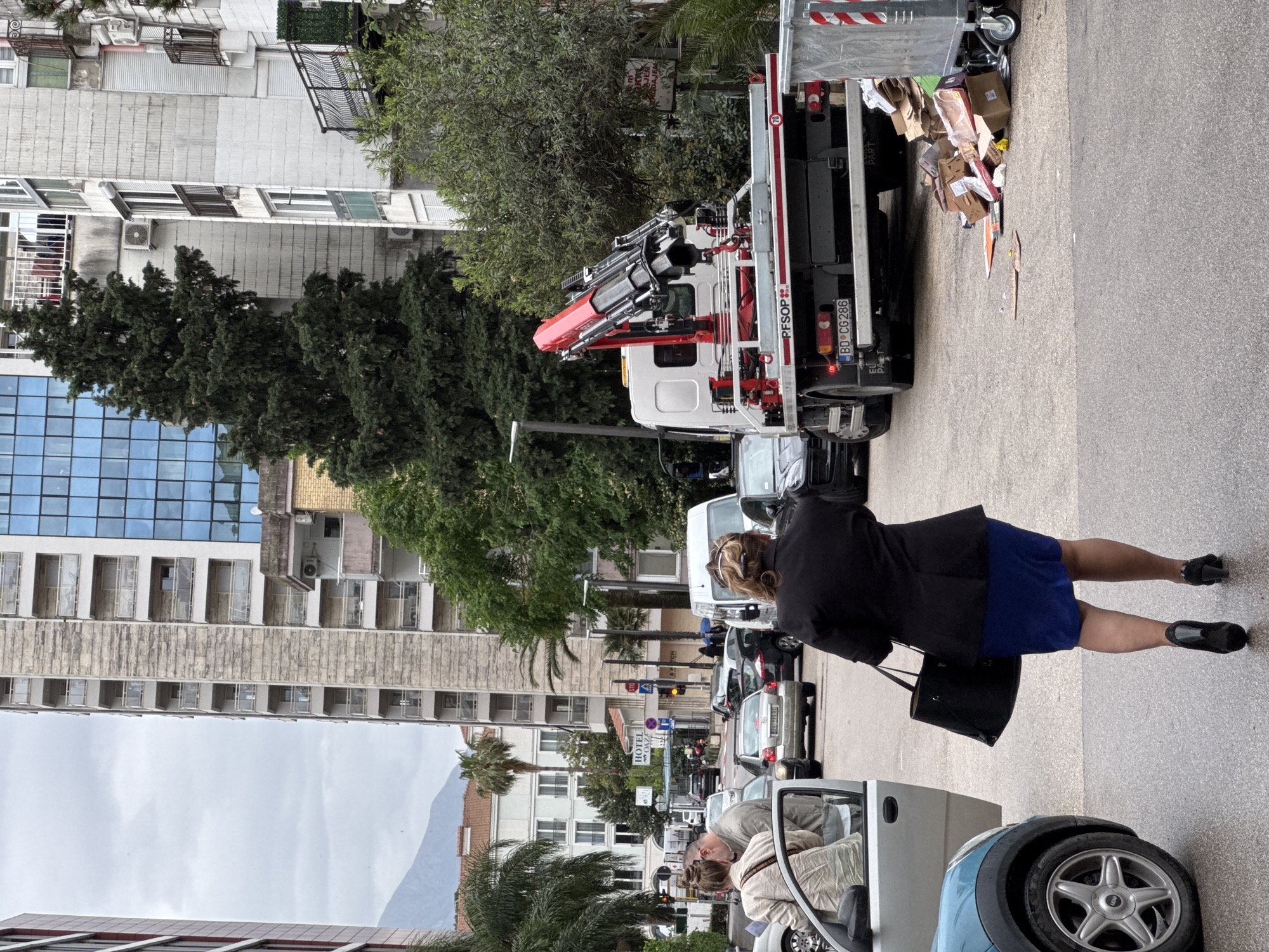 This image captures a candid street scene in Budva, Montenegro, during an overcast daytime. The photograph is oriented sideways, requiring a mental rotation to view correctly.
The scene features a mix of urban architecture, lush greenery, and human activity on a street. In the foreground, a light blue car is parked, with two individuals interacting: one person is seated inside, while another leans into the car from the driver's side. To their right, a woman in a black jacket, blue skirt, and black shoes walks away from the camera, carrying a black handbag.
Further down the street, a white truck equipped with a red hydraulic crane is prominently parked. Its bed is filled with various cardboard boxes and what appears to be general refuse. The truck bears the license plate "BD CG286," indicating Budva, Montenegro, and the text "PFSOP" is visible on its side. Adjacent to the truck, scattered waste is visible on the asphalt.
Buildings line both sides of the street. On the left, a modern building with a large blue glass facade stands alongside multi-story residential or commercial structures featuring balconies, air conditioning units, and roller shutters. On the right, older buildings are interspersed with trees, including palm trees. A sign for "HOTEL" is visible in the mid-ground, partially obscured. Traffic signs, including a "STOP" sign and a traffic light displaying yellow, are also present. In the far distance, a mountain range is visible under a cloudy sky.
The overall impression is one of everyday life in a bustling coastal town, with people, vehicles, and commercial activities coexisting.