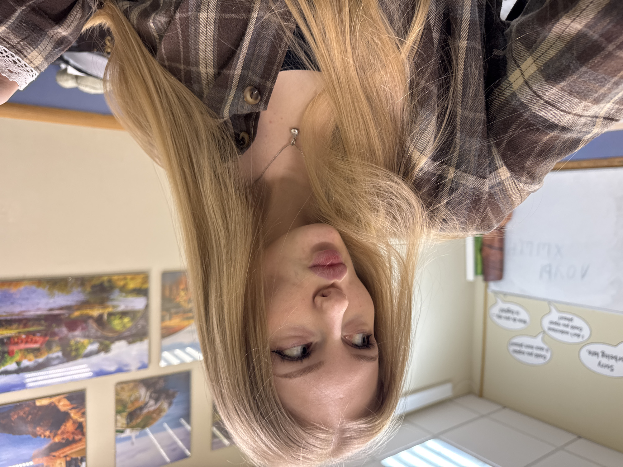 An image, rotated 90 degrees clockwise, captures a young woman from a low-angle perspective, making her appear upside down. She has long, straight blonde hair that cascades around her shoulders. Her eyes are light, possibly blue or green, and she is looking upwards and to her left, away from the camera, with her lips slightly pursed in what appears to be a pensive or playful expression.
She is wearing a plaid shirt in shades of brown, beige, and dark grey. A delicate silver necklace with a small, clear gemstone pendant is visible on her neck. A white lace cuff of a garment is peeking out from under her plaid sleeve on the left side of the frame.
The background suggests an indoor setting, possibly a classroom or common area. On the wall behind her, several framed pictures depicting natural landscapes are displayed. To her right, a whiteboard is visible, with faint, inverted writing that includes what looks like "AKOV" and possibly "MMXX". Below the whiteboard, a series of speech bubble-shaped cutouts are affixed to the wall. One clearly visible, inverted bubble reads: "Sorry for being late." Other text on the bubbles is partially obscured or indistinct due to the angle and inversion, but one seems to begin with "I don't remember...". The floor appears to be tiled, and reflections of overhead strip lighting are visible on surfaces.