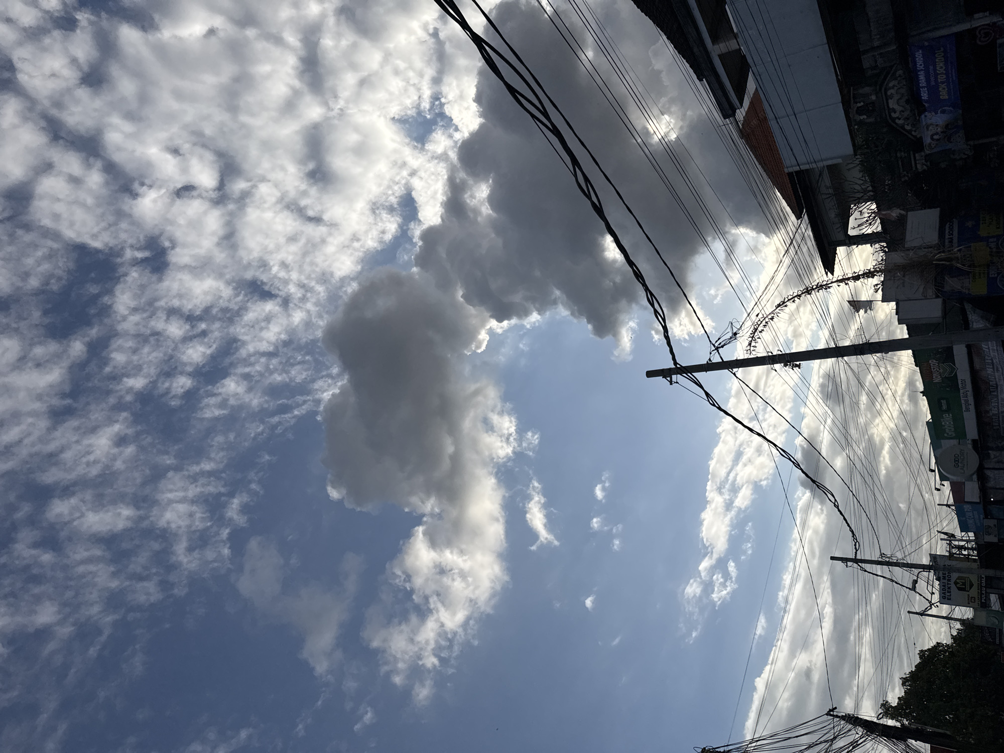 This image captures a street scene in Denpasar, Indonesia, dominated by a dramatic sky filled with a mix of dark storm clouds and brighter, wispy cirrus clouds against a blue sky. Numerous utility wires crisscross the frame, supported by utility poles, indicating an urban or semi-urban environment. In the foreground and mid-ground, buildings with traditional Indonesian architectural elements, such as terracotta tiled roofs, are visible. Several signs for businesses are present, including "Nonstop," "Gadai ME ELEKTRONIK," "GrabBike Hebat," and "ARIE RAMA SCHOOL Welcome to BACK TO SCHOOL." No people are clearly discernible in the image. The overall impression is of a bustling street under a dynamic and possibly changing weather condition.