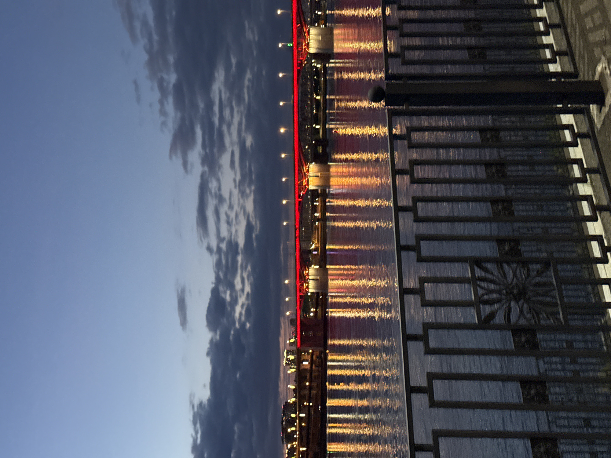 The image captures a bridge over the Yenisey River in Krasnoyarsk, Russia, at twilight. The view is from a pedestrian walkway with an ornate, dark metal railing featuring stylized flower-like designs. The river reflects the warm, golden light from the bridge and surrounding city lights. A line of red lights illuminates the edge of the bridge. The sky transitions from a clear, light blue to darker blue with cloudy patches. No people are visible.