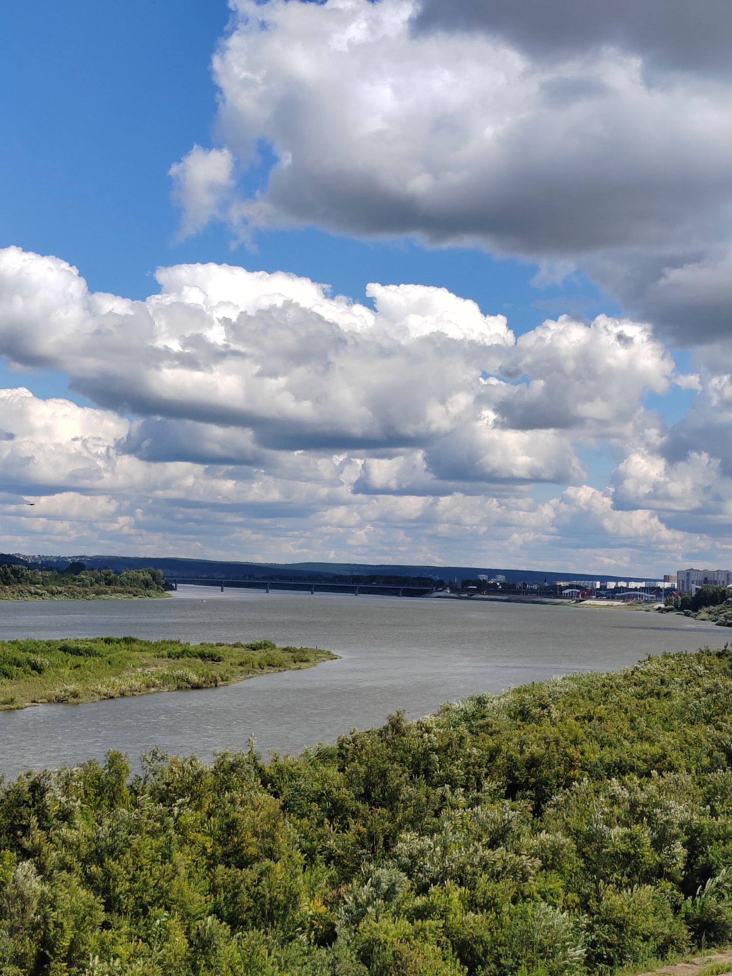The image depicts a panoramic view of the Tom River in Kemerovo, Russia, on a bright, cloudy day. In the foreground, lush green foliage and trees cover the landscape, leading down to the river. The river itself is wide and calm, with a gentle curve. Across the river, the opposite bank is lined with trees and further back, buildings and structures indicative of a city are visible. A long bridge spans the river in the distance, connecting the two sides. The sky is a vibrant blue, filled with numerous large, fluffy white and grey clouds, creating a dramatic and dynamic atmosphere. The scene conveys a sense of natural beauty combined with urban development.