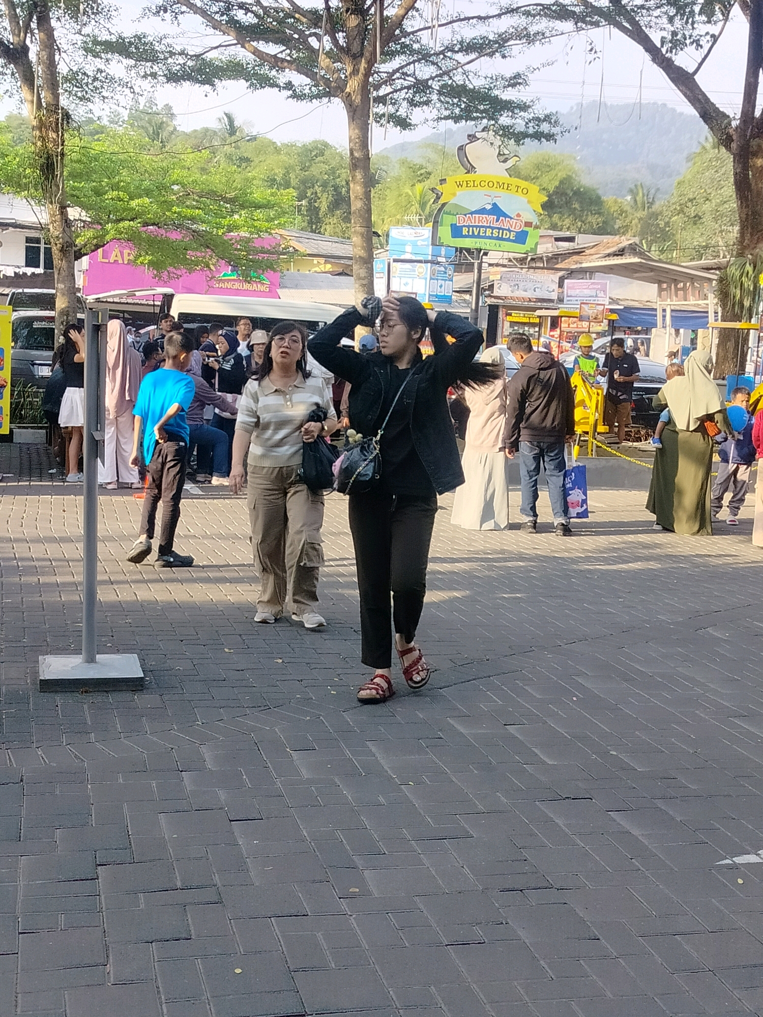 A group of people are standing on a paved walkway in front of buildings. A sign that says "WELCOME TO DAIRYLAND RIVERSIDE - PUNCAK" is visible in the background. The walkway is made of dark gray brick pavers arranged in a pattern. Trees with green foliage are in the background, and the sky is bright and overcast. In the foreground, a woman with dark hair is walking towards the viewer. She is wearing a black jacket, black pants, and red sandals. She is holding her hands up to her head. Behind her, to the left, a woman in a striped shirt and beige pants is standing and looking to the left. She is holding a black bag. To the left of the woman in the striped shirt, a young boy in a blue shirt and black pants is walking away from the viewer. Other people are visible in the background, some standing in front of buildings and others walking. The overall scene appears to be an outdoor marketplace or tourist attraction.