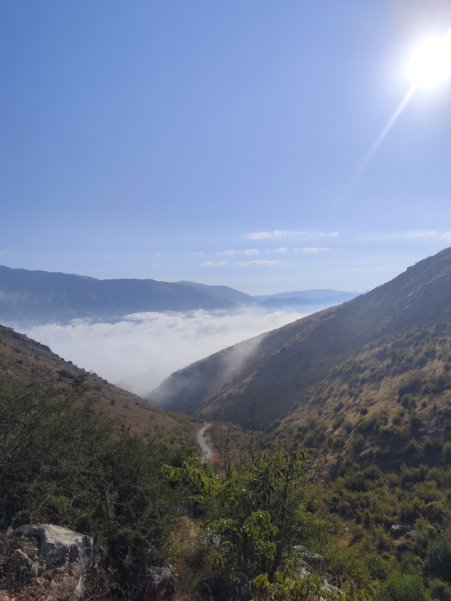 The image depicts a mountainous landscape, likely in Dropull i Sipërm, Albania, on a clear, sunny day. In the foreground, dry, rocky terrain is covered with sparse, brown vegetation and scattered green bushes. A winding road is visible in the mid-ground, leading into a valley filled with a thick layer of white clouds or fog. Beyond the fog, several layers of mountains recede into the distance, with the furthest peaks appearing hazy blue. The bright sun is positioned in the upper right corner of the frame, casting a glare and creating lens flares. The sky is a vibrant, clear blue with a few wispy white clouds. There are no people or animals visible in the scene. The overall impression is one of a remote, natural environment bathed in sunlight.
