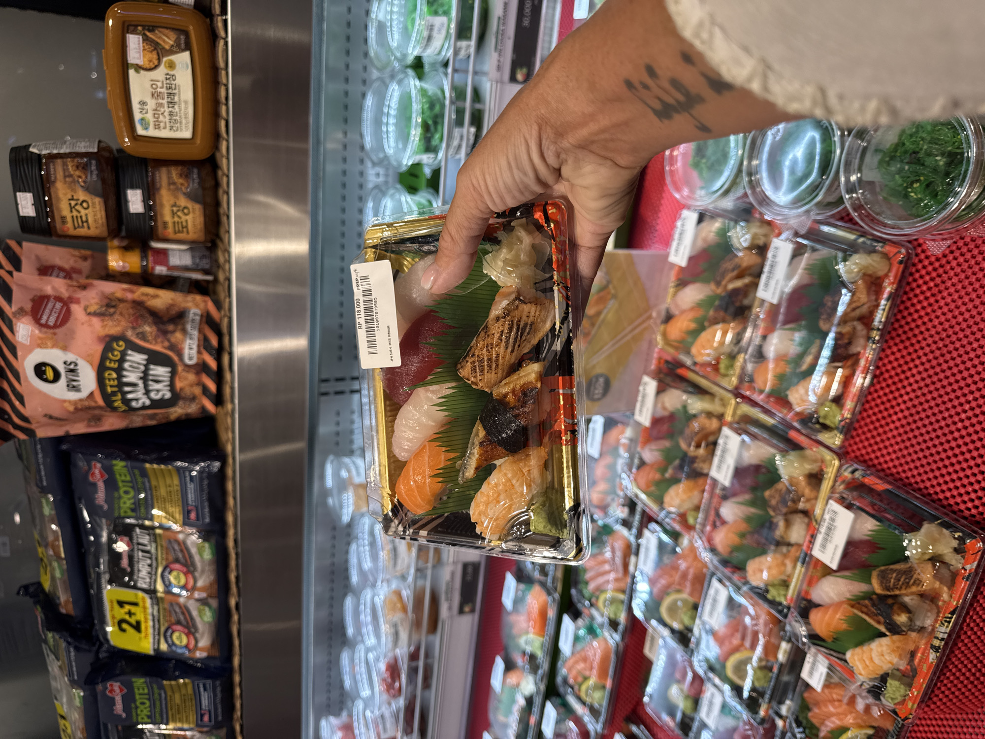 An eye-level indoor shot shows a person's hand holding a packaged sushi tray in a supermarket's refrigerated section in Kuta Selatan, Indonesia. The hand, with a tattoo on the wrist, is centered in the frame, presenting the clear plastic container of assorted sushi to the viewer. The sushi tray contains various types of nigiri and possibly some maki, with ginger and wasabi visible, presented on a green leaf. A white label on the package displays "RP 118.000", "JPN SUSHI MIXS MEDIUM", and a barcode with the number "101087079505".
In the background, more identical sushi packages are neatly arranged on a red, textured mat, suggesting a display area. To the left, several other food products are stacked on shelves. These include bags of "IRVINS SALTED EGG SALMON SKIN" snacks, and packages of "RUMPUT LAUT PROTEIN" seaweed snacks, some marked with "2+1". Further left, jars of "토장" (Doenjang), a Korean fermented soybean paste, from the "신송" (Sinsong) brand are visible, with text "짠맛을 줄인 건강한 재래된장" (Reduced salt, healthy traditional doenjang). To the right and behind the sushi display, small clear plastic containers of what appears to be "HURA WAKAME" (seaweed salad) are stocked, with one visible price tag of "30,000". The overall scene depicts a well-stocked grocery store aisle, likely a dedicated section for international or prepared foods.