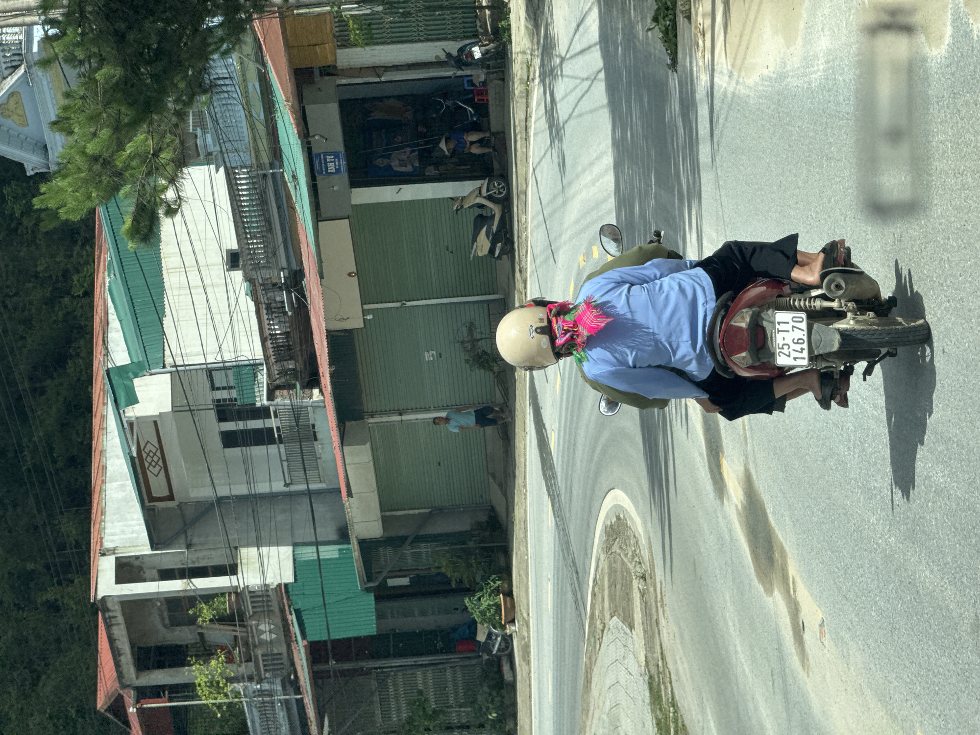The image depicts a daytime street scene in Púng Luông, Vietnam, observed from a vehicle. In the immediate foreground, a red Honda motorcycle with license plate "25-T1 146.70" travels away from the viewer on an asphalt road. Two individuals occupy the motorcycle. The driver wears a light blue long-sleeved shirt, black wide-leg pants, and black sandals. A cream-colored motorcycle helmet covers their head, and a vibrant pink, green, and white patterned scarf is visible at the back of their neck. A second passenger is partially obscured behind the driver.
The asphalt road, marked with a faded yellow line and minor stains, curves gently to the left. To the right of the road, a sidewalk transitions to a row of multi-story buildings. These structures exhibit varied architectural styles, featuring elements such as red and green corrugated metal roofs, white and light-colored facades, balconies with railings, and numerous exposed utility wires traversing the scene. Several storefronts are visible at ground level; most have closed green roll-down shutters.
In the midground, a man in a light blue shirt and dark shorts stands idly near a closed green-shuttered shop. Further along, an open storefront with a blue "ANH TƯ" sign reveals a person wearing a traditional conical hat, seated on a low stool. Two parked scooters, one light-colored and one dark, are visible near this open shop.
The background is dominated by dense, dark green foliage covering a hillside, suggesting a mountainous or heavily vegetated environment. A partial view of a white building with an ornate roofline is visible through the trees in the upper right. Strong shadows indicate bright sunlight.